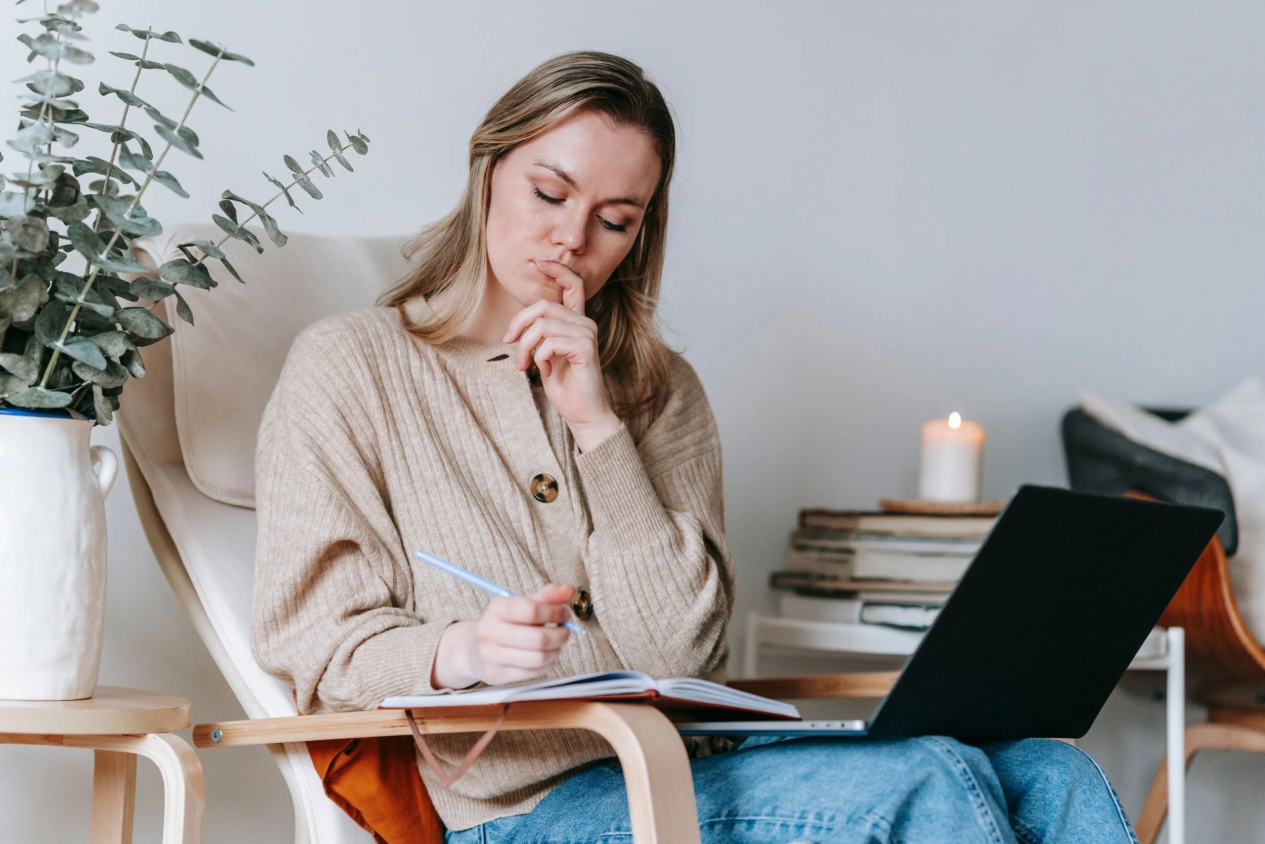 A young woman with blonde hair sitting on a wooden chair, holding a pen and writing in a notebook, with a laptop in front of her. She appears thoughtful, with her hand near her mouth, in a cozy indoor setting with a candle, a plant, and a stack of books in the background.