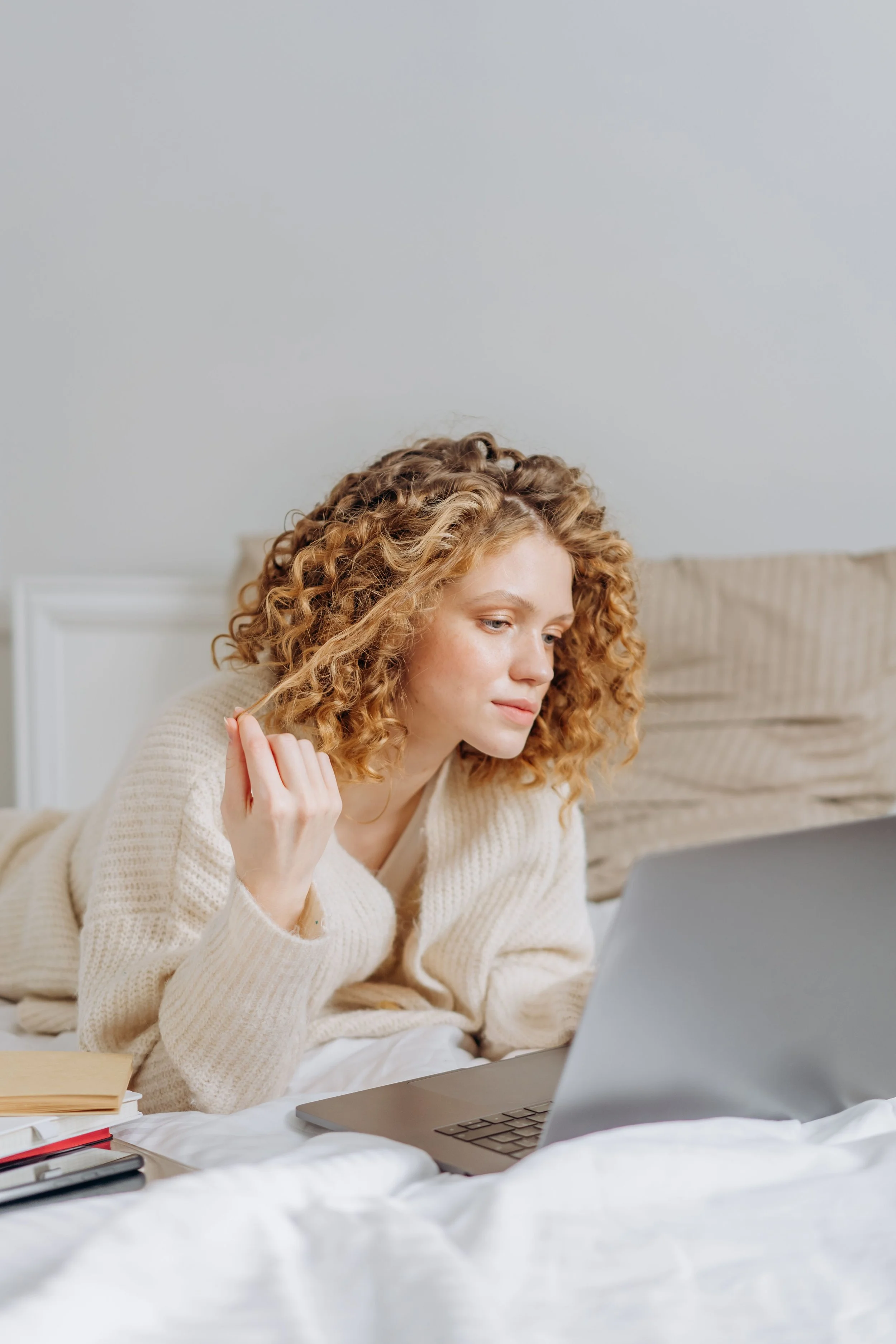 A woman with curly blonde hair, wearing a beige sweater, sitting on a bed with a laptop in front of her, in a bedroom with white walls and a beige headboard.