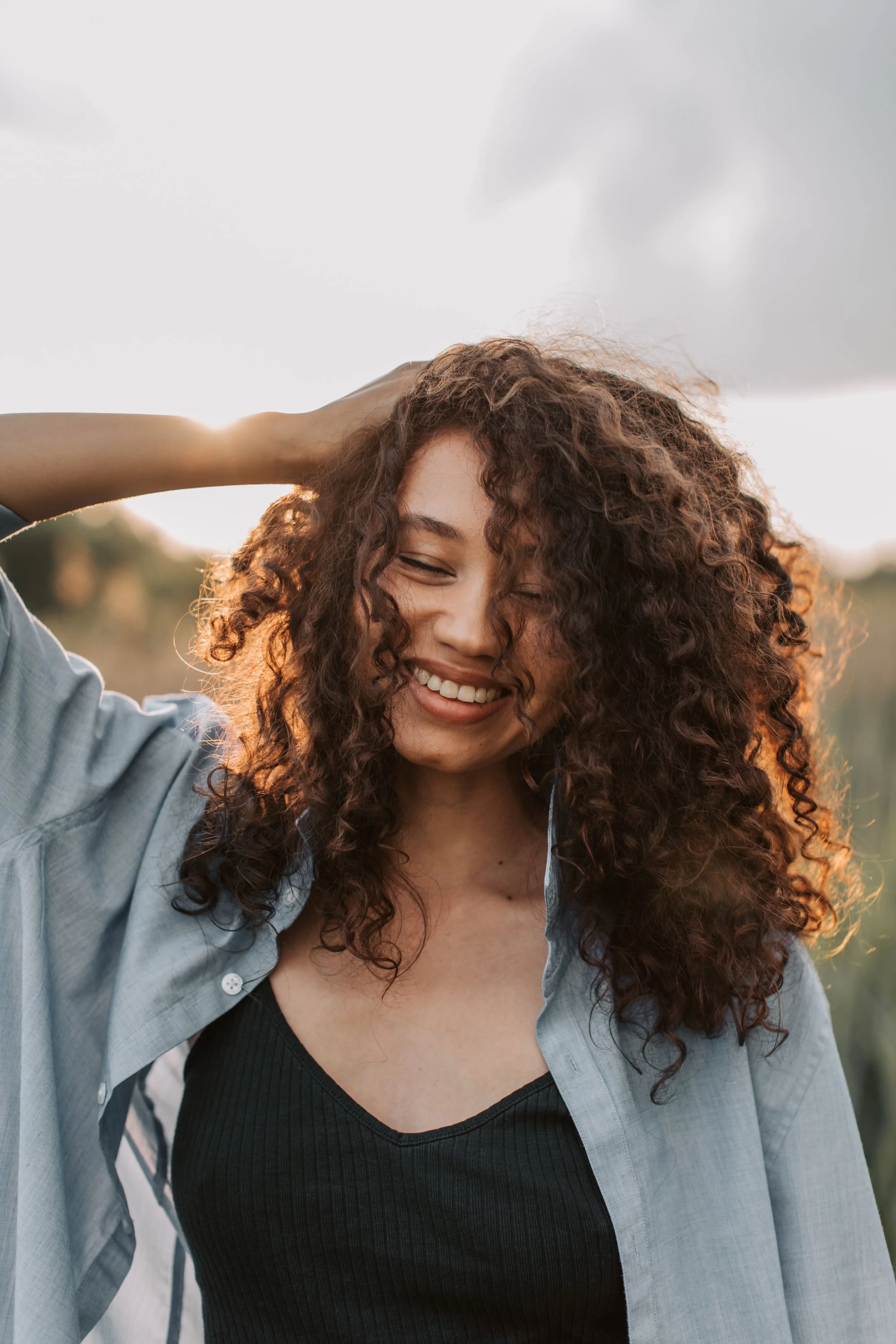 A woman with curly hair smiling outdoors, wearing a blue shirt and a black top, with her hand on her head, during sunset or sunrise.
