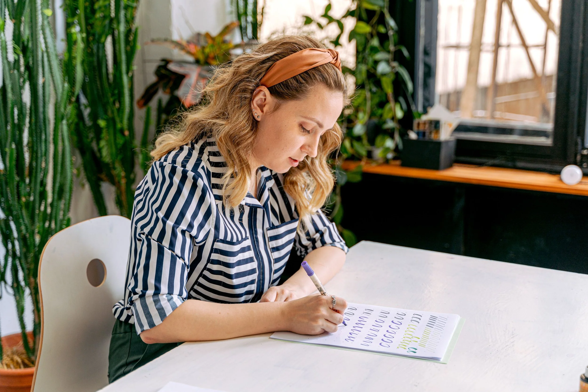 A woman with wavy blonde hair wearing a tan headband and a black and white striped shirt is sitting at a white table, writing in a notebook with colorful lettering on the pages. There are green plants behind her and a black window frame with a view of a garden.