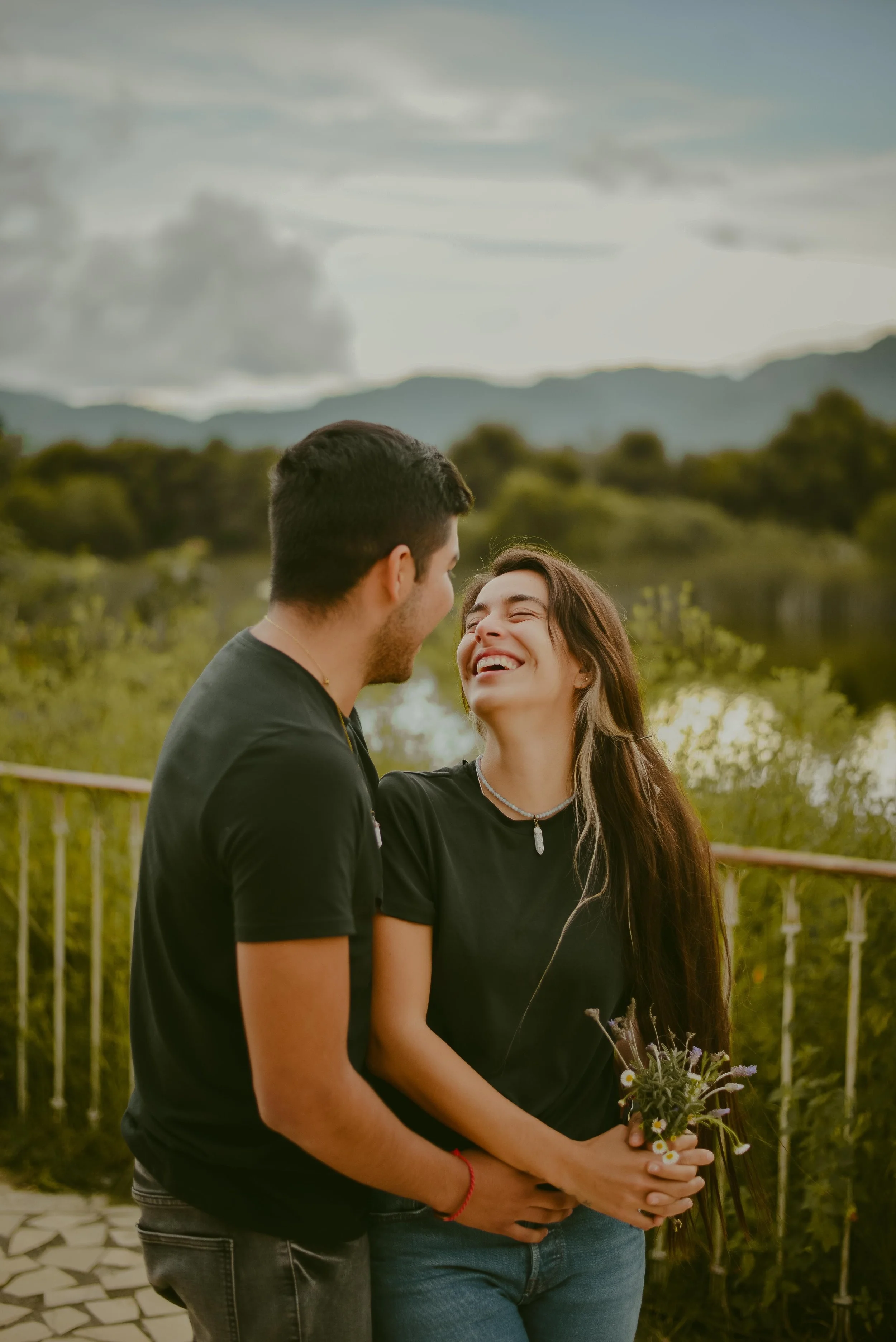 A couple standing close together outdoors, smiling and looking at each other, with a scenic background of a lake, trees, and mountains.