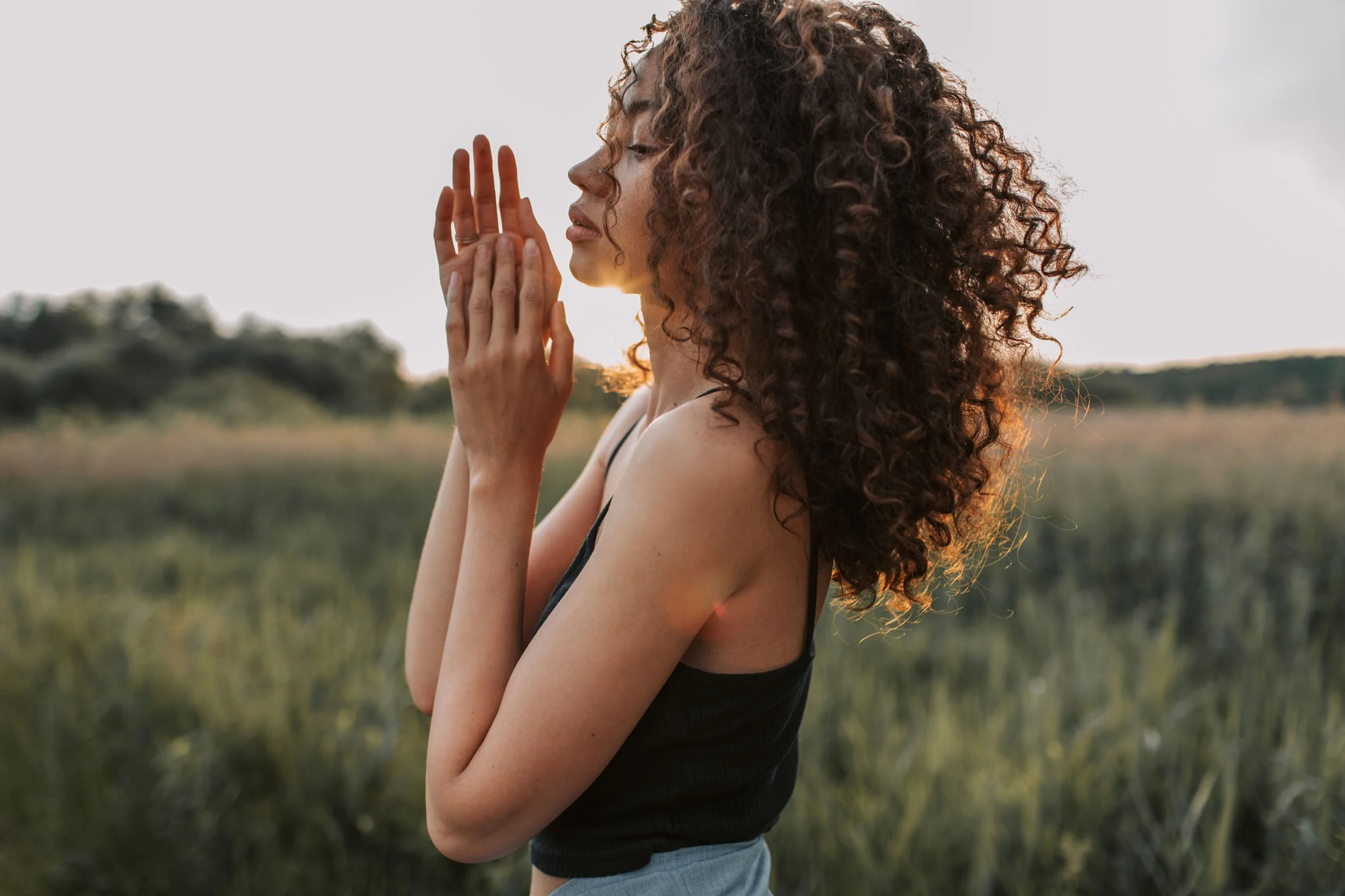 A woman with curly hair standing outdoors in a field, with her hands near her face, wearing a tank top and standing sideways.