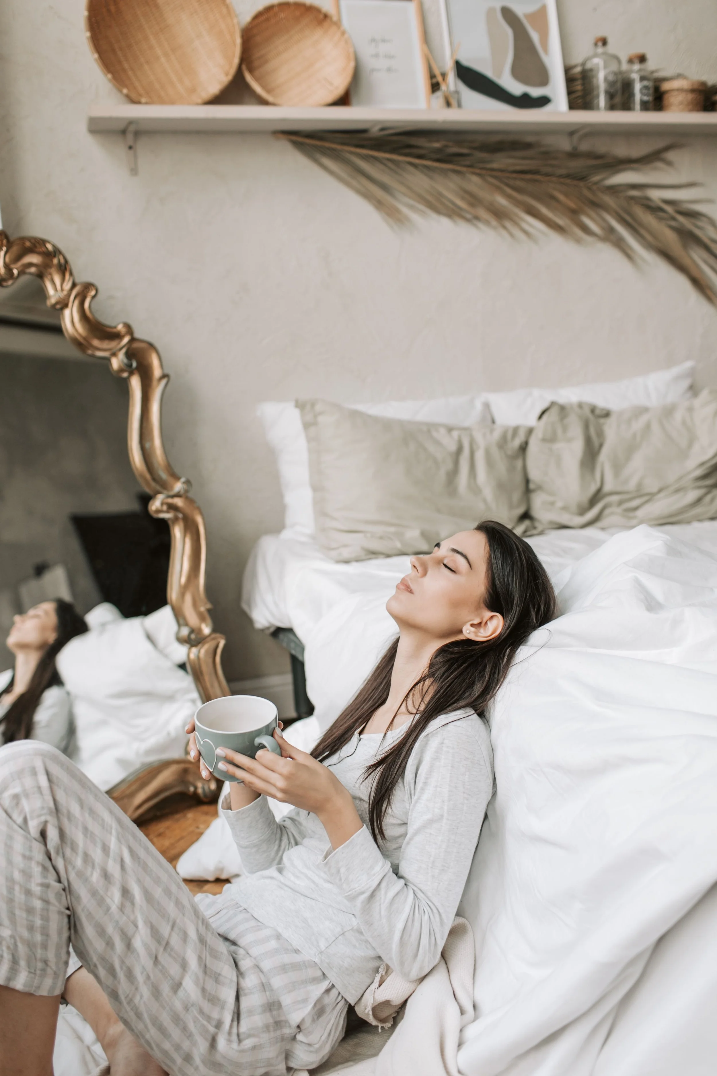 Two women relaxing in bed, one is holding a mug and the other is resting with eyes closed. The bed has white bedding and headboards. There is a decorative mirror, a mirror with a gold frame, and shelves with decor items above.