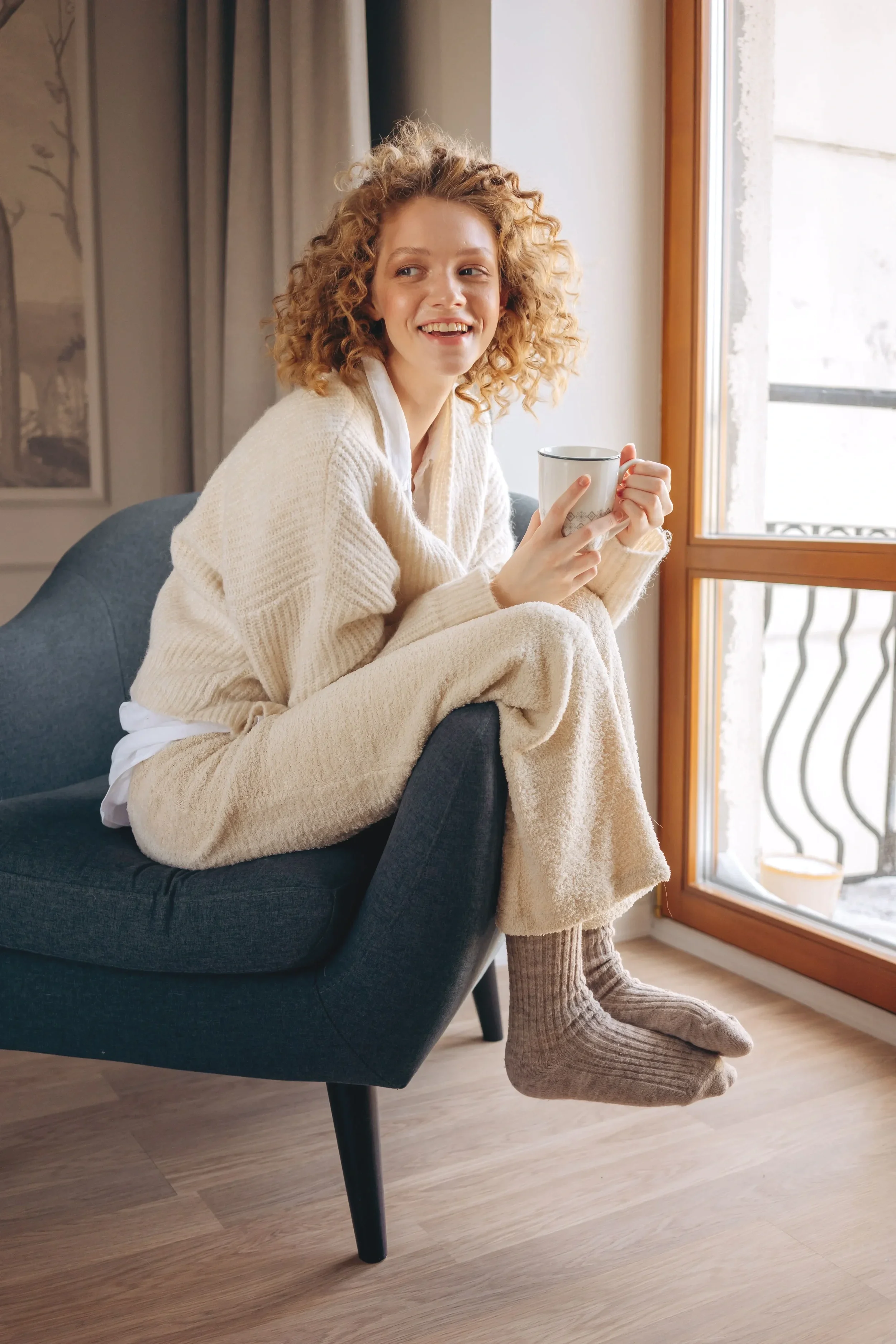 A woman with curly red hair sitting on a modern black armchair by a large window, holding a coffee mug and smiling, dressed in cozy beige and white clothes, with slippers, in a warmly lit room.