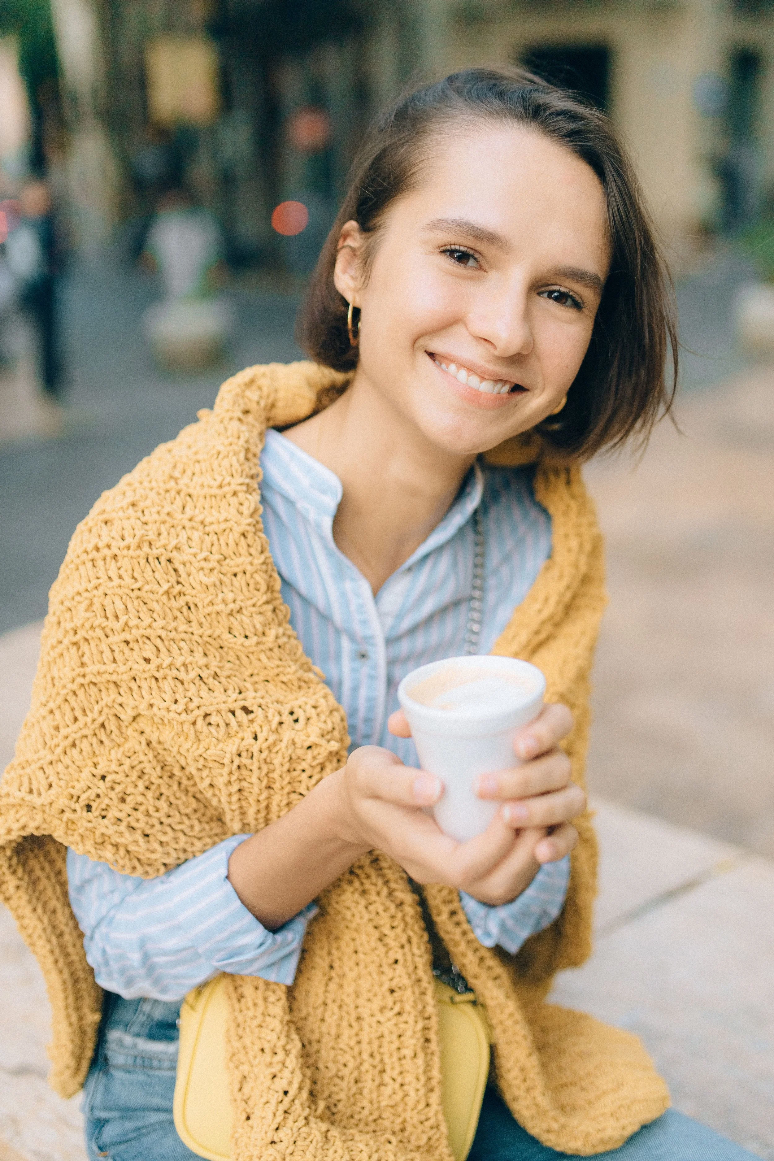 A young woman with short brown hair smiling and holding a styrofoam cup with a beverage in an outdoor urban setting.