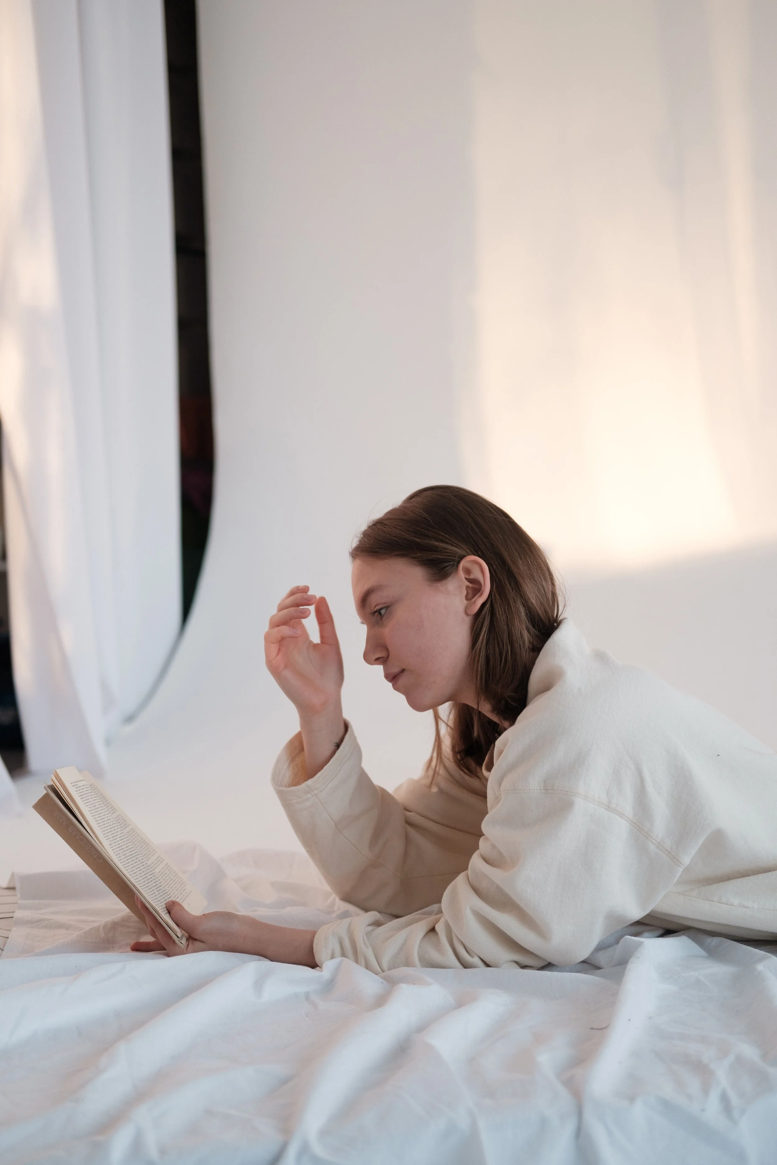 A woman with brown hair sitting and reading a book on a bed with white sheets, holding her head with her left hand. She is wearing a cream-colored hoodie and appears to be in a softly lit room with white curtains.