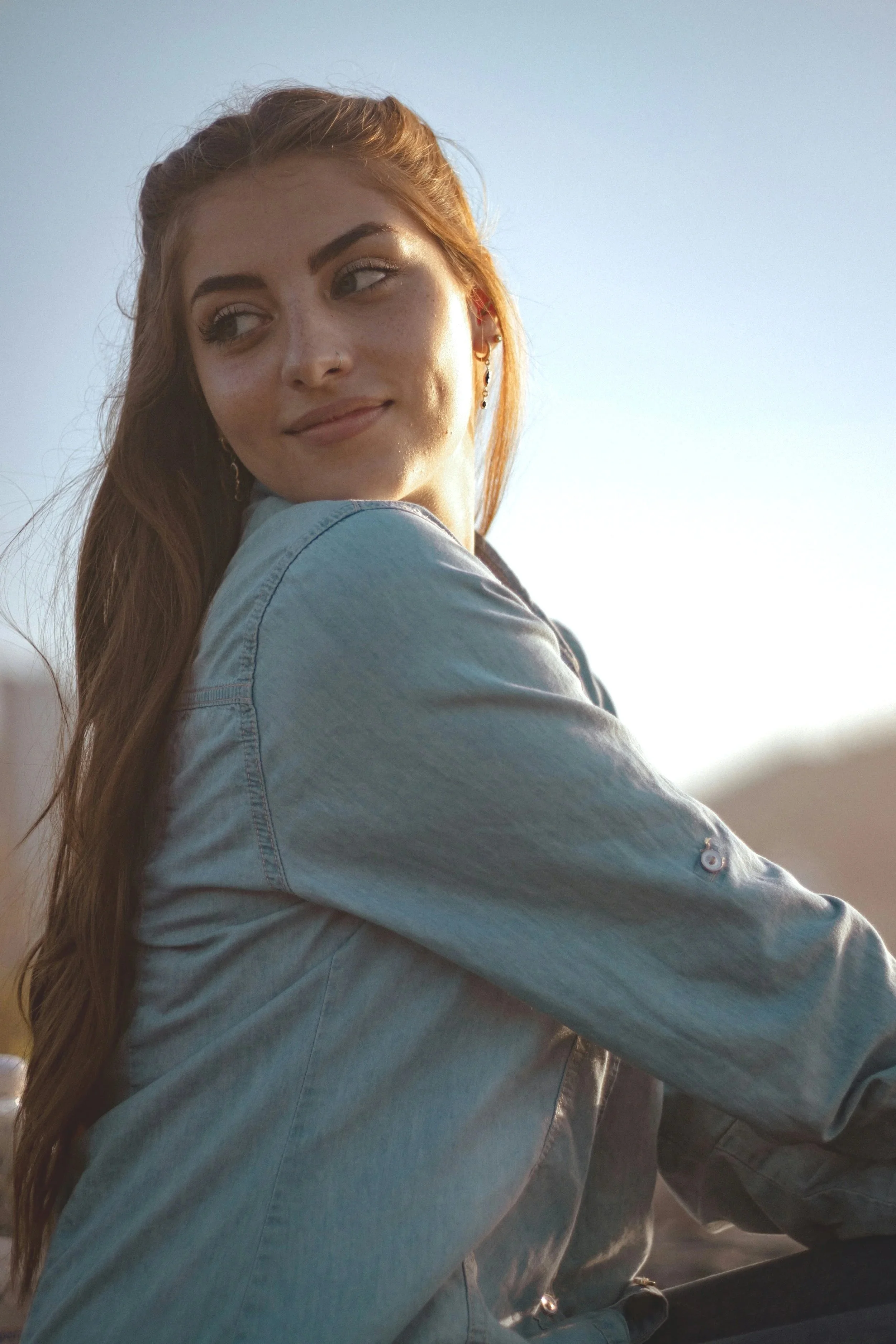 Close-up of a young woman with long, reddish hair, wearing a denim jacket, looking over her shoulder with a soft smile, outdoors during golden hour sunlight.
