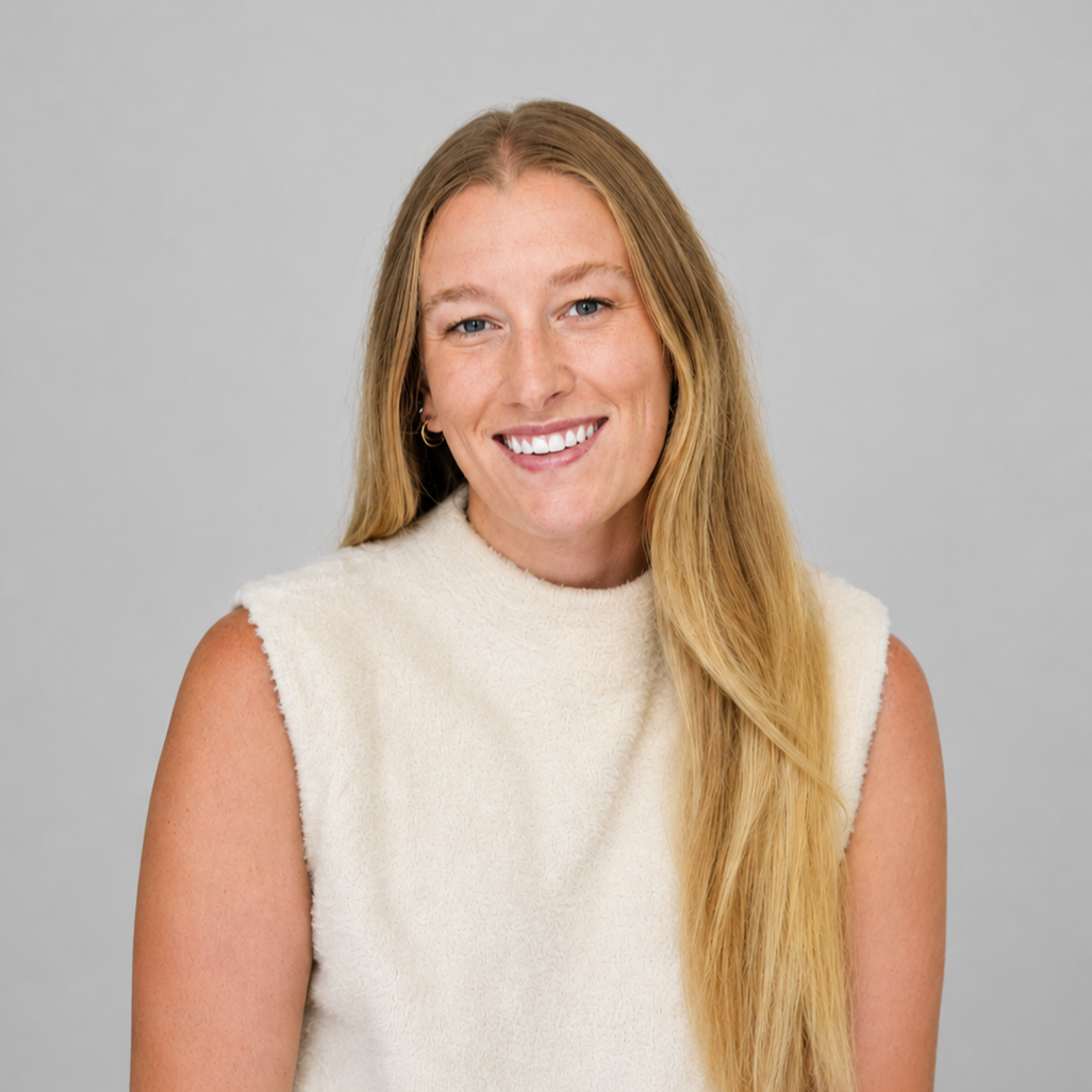 A woman with long blonde hair, wearing a sleeveless cream-colored top, smiling against a plain gray background.