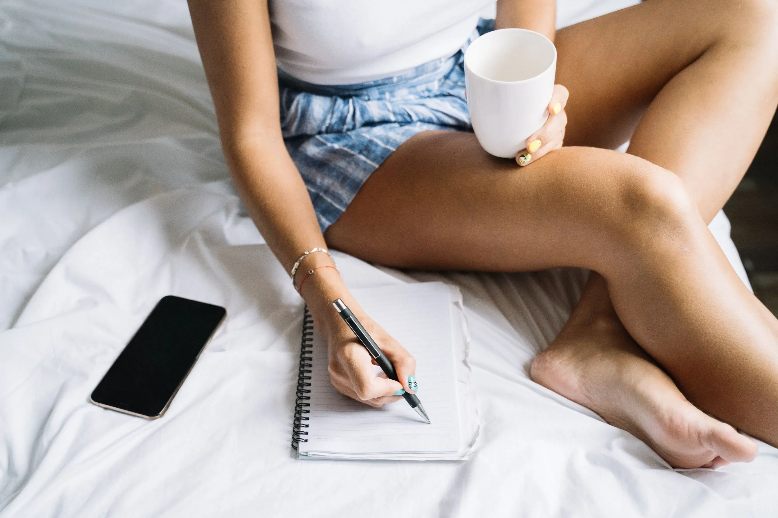 Individual sitting in bed reflecting in her journal while holding a coffee