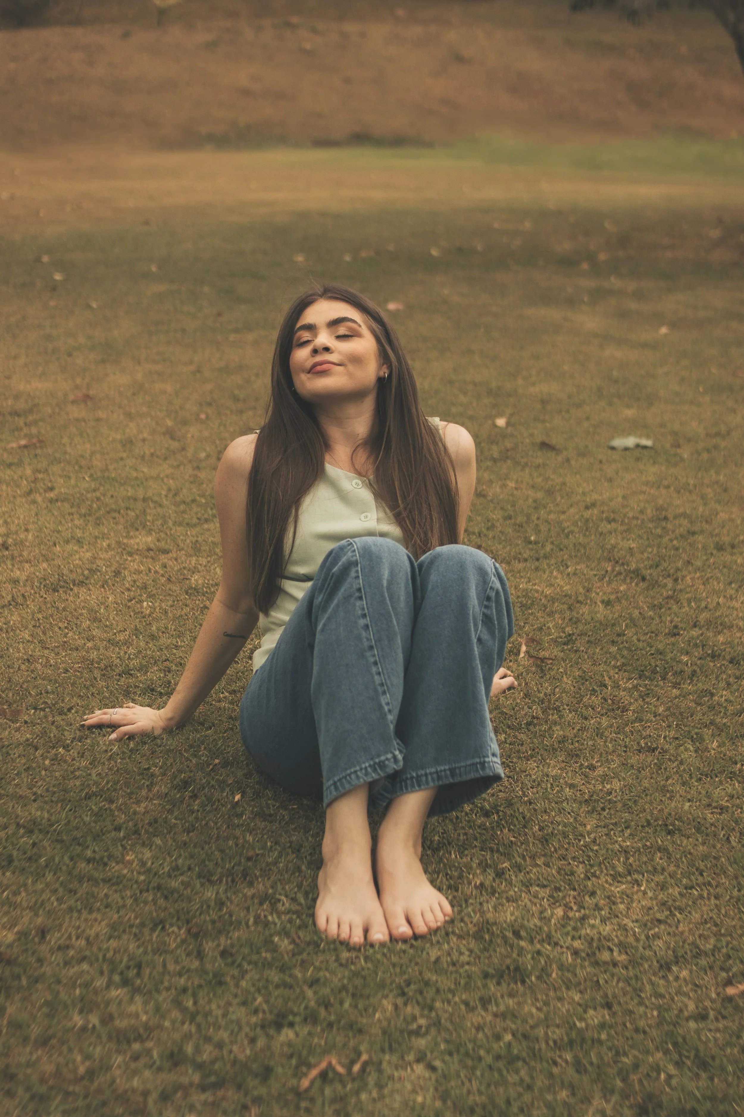 A young woman with long dark hair and closed eyes sitting on the grass with her legs crossed and relaxed, in a natural outdoor setting.