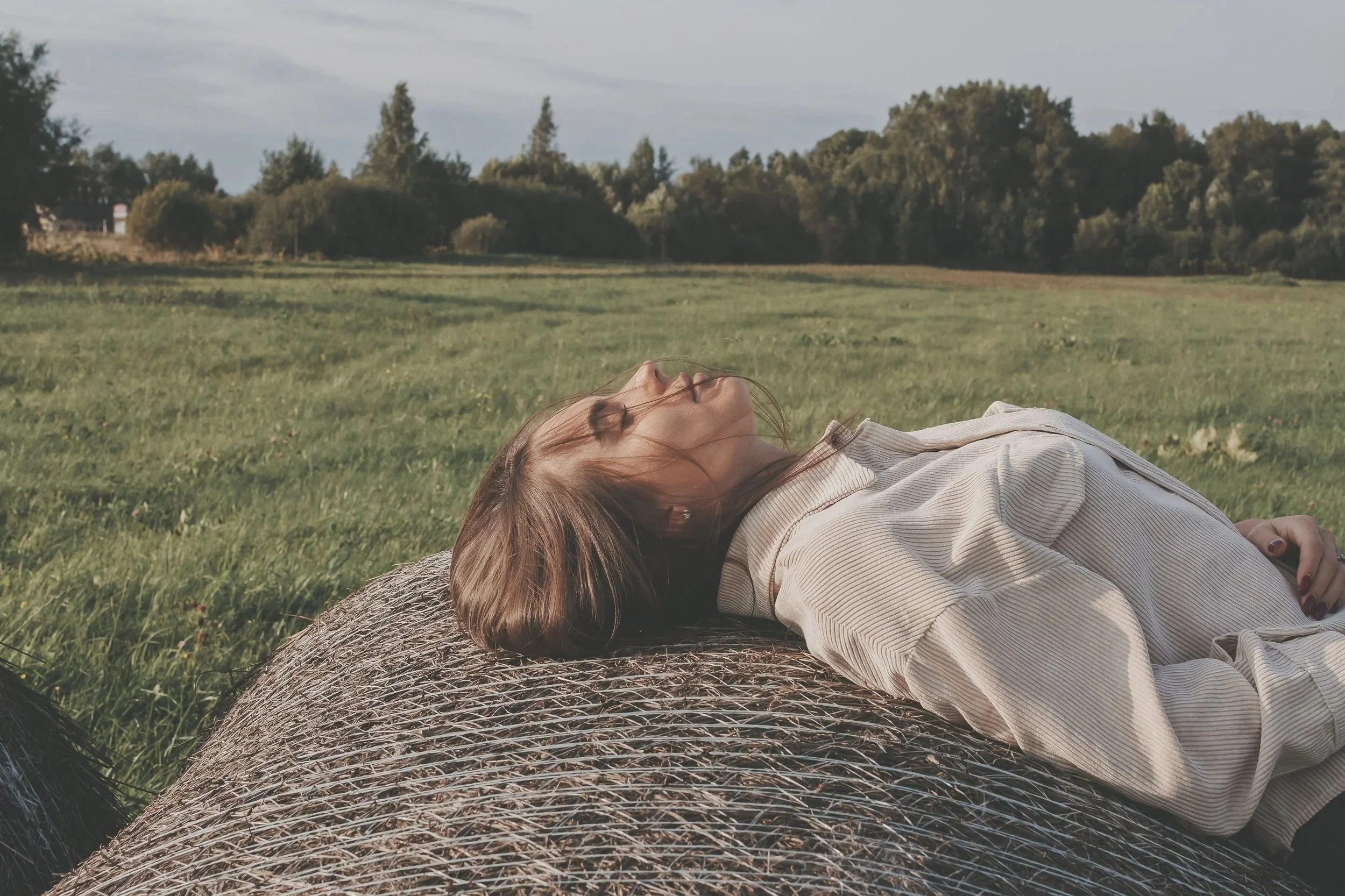 A woman lying on a hay bale in a grassy field with trees and a cloudy sky in the background