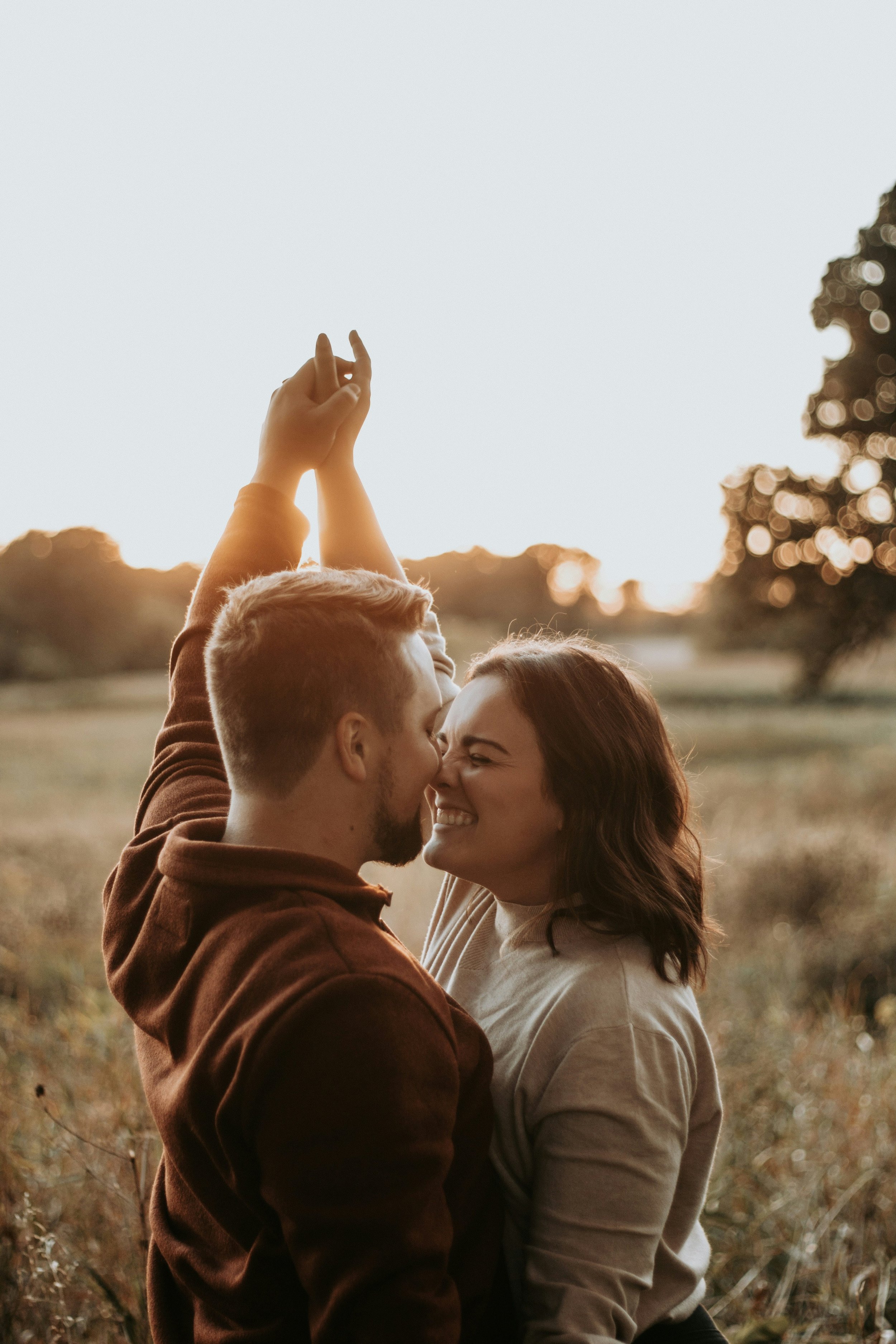 A couple stands close together outdoors during sunset, with a man and woman touching foreheads and smiling, surrounded by nature and trees with blurred bokeh lights in the background.