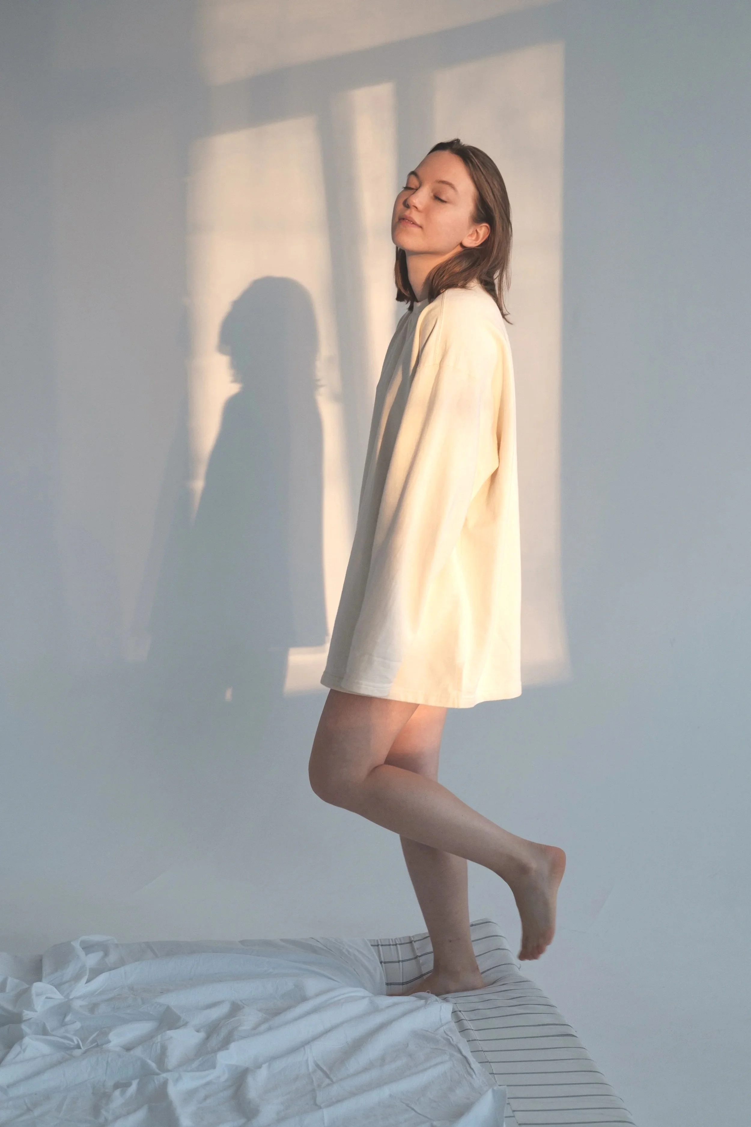 A woman with shoulder-length brown hair wearing a cream-colored long-sleeve shirt standing on a bed, with one foot on a striped pillow and the other foot lifted, casting a shadow on the light blue wall behind her.
