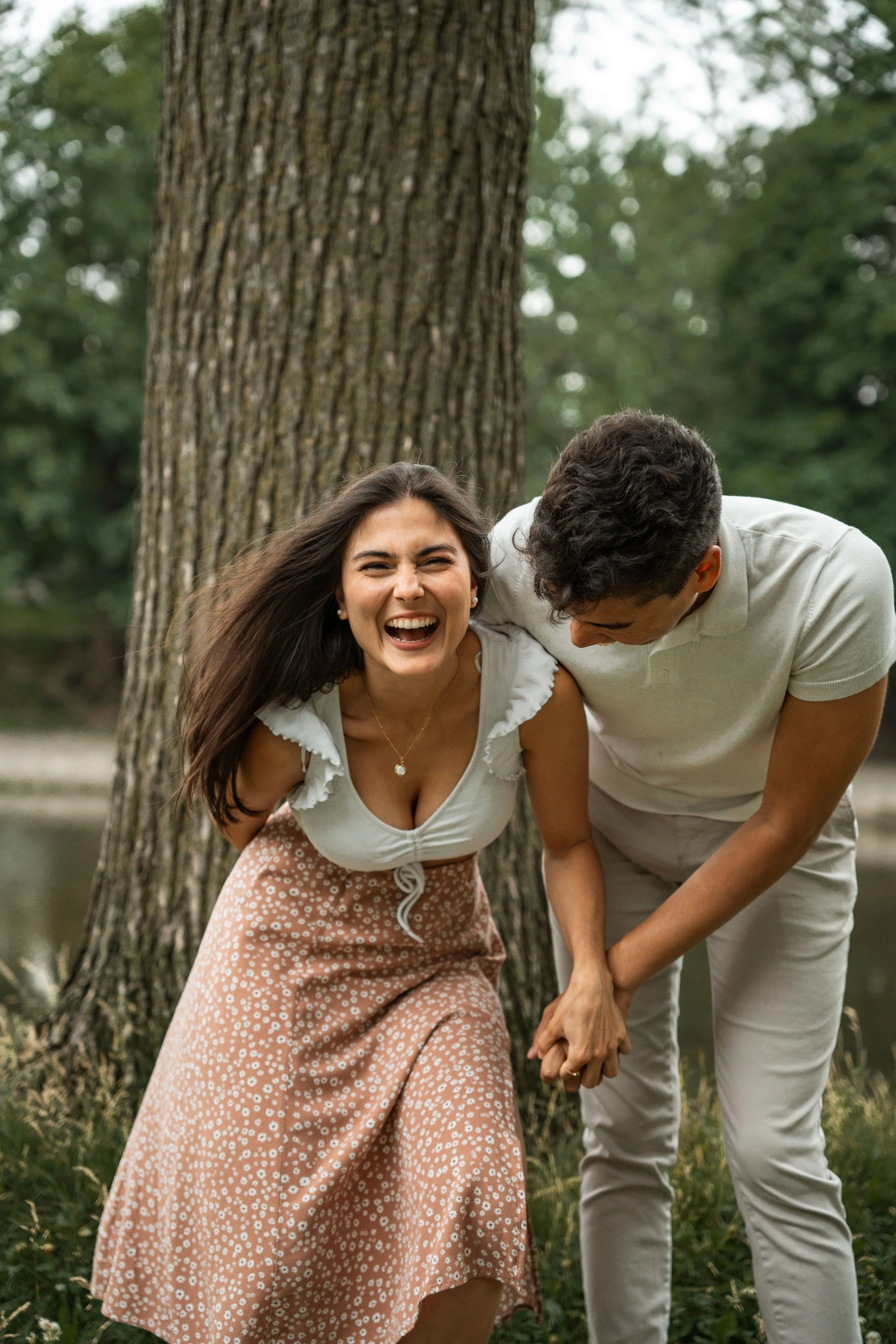 A young woman and a young man are outside near a large tree, holding hands and laughing together. They are in a natural setting with greenery and a lake in the background.