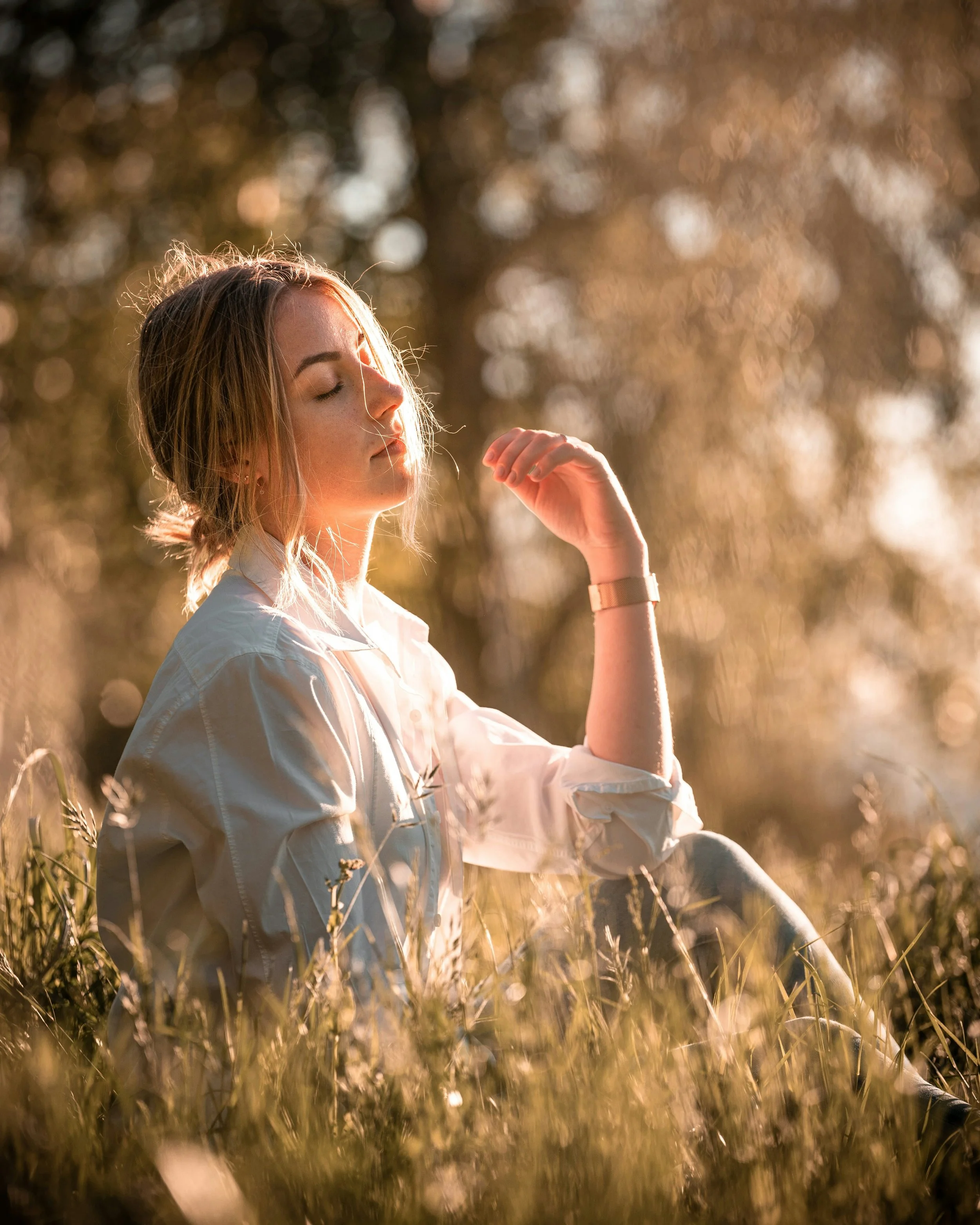 A woman with closed eyes sitting in a field of tall grass, bathed in warm sunlight with a blurred background of trees.