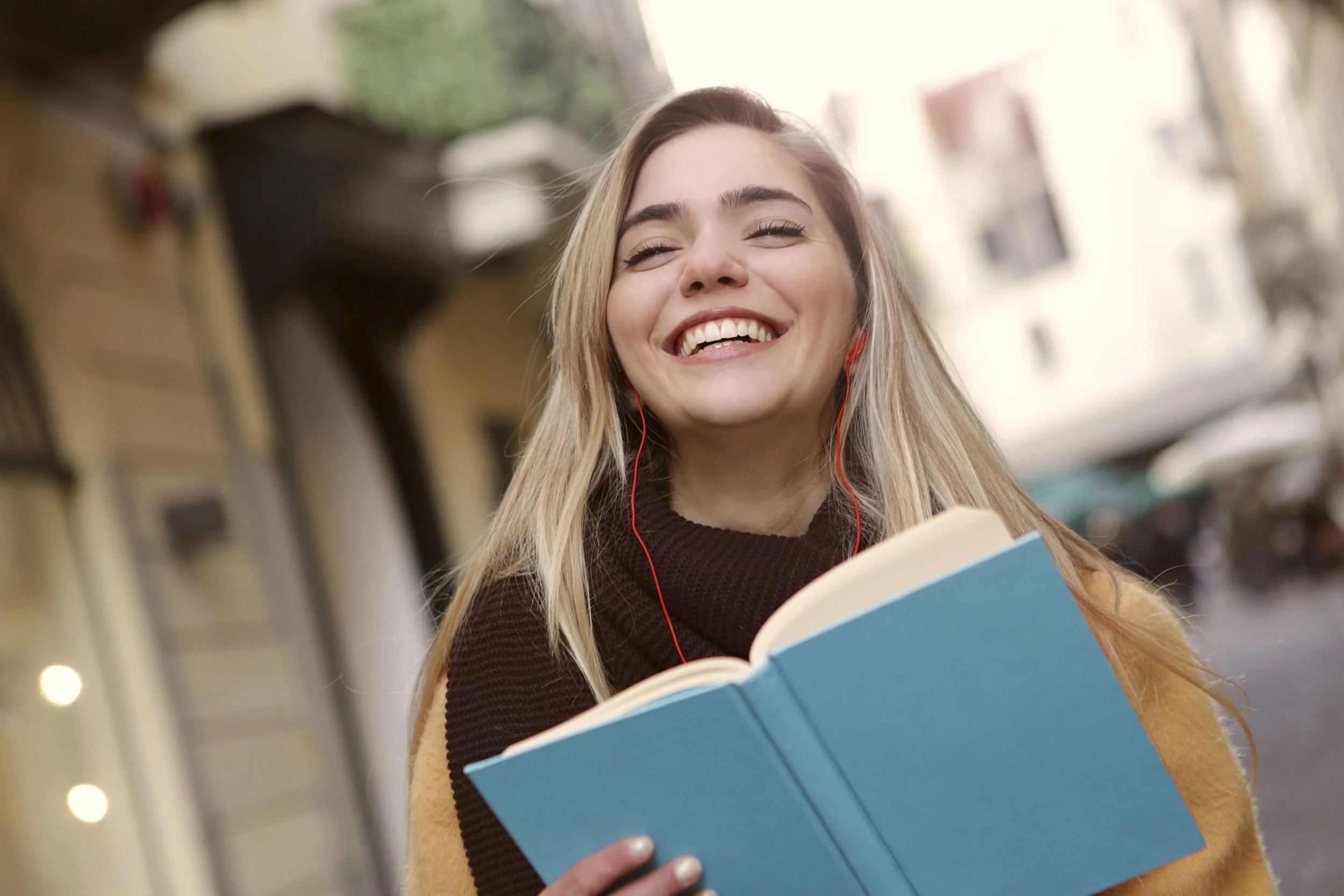 A young woman with long blonde hair wearing a beige coat and brown sweater, smiling with eyes closed, holding a blue book and listening to red earbuds, standing in a store aisle.