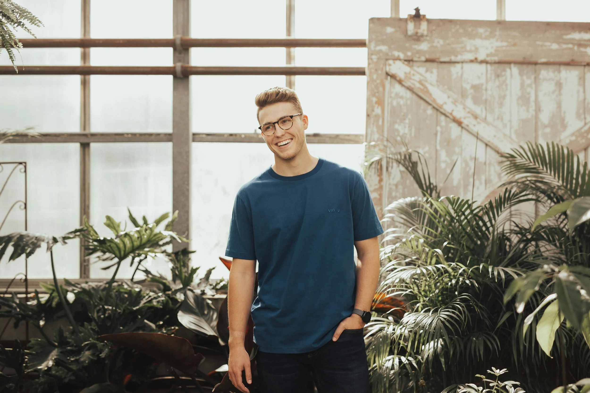 A young man wearing glasses and a blue t-shirt smiling in a greenhouse surrounded by lush green plants.