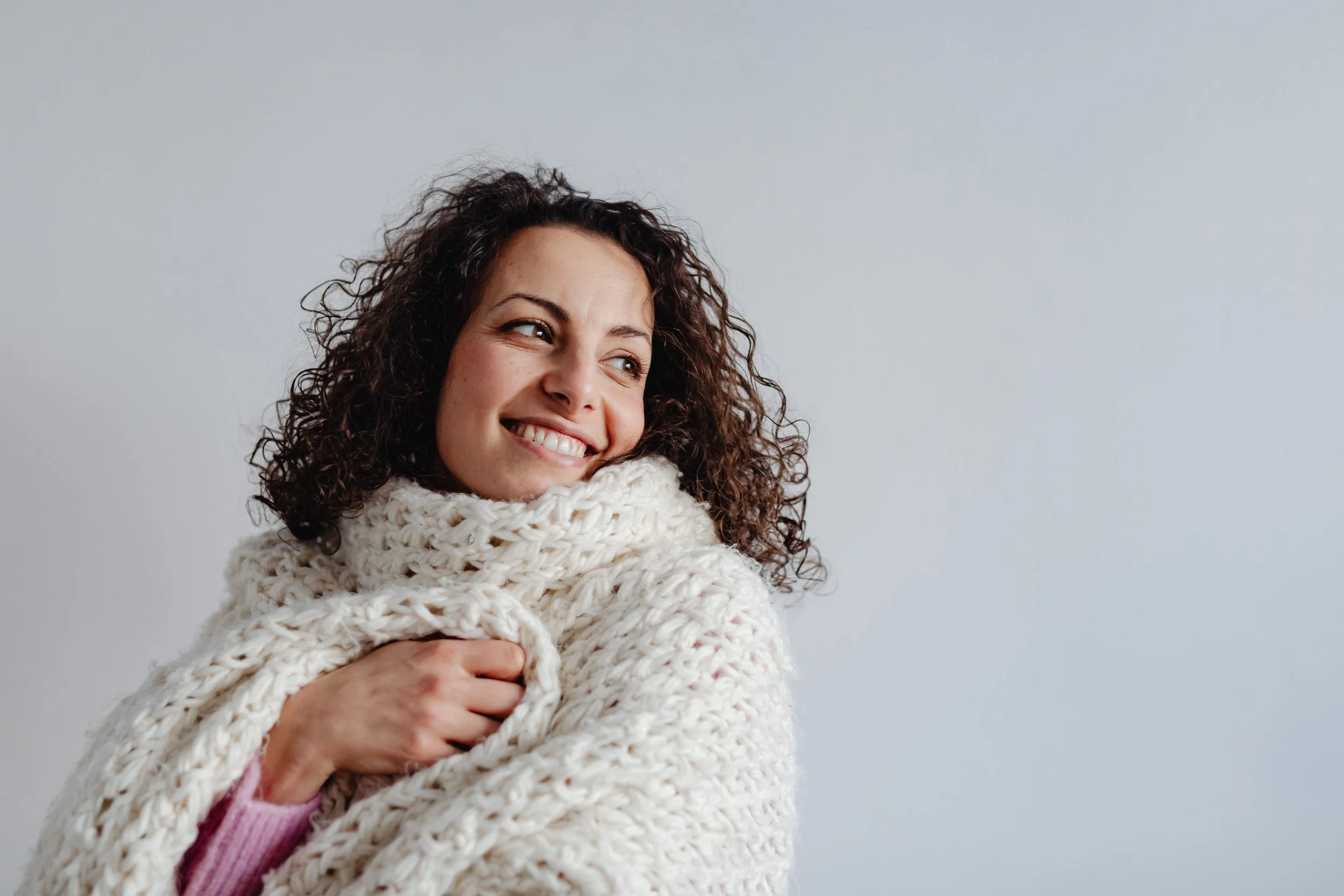 A woman with curly hair smiling and hugging herself in a chunky knit sweater against a plain grey background.