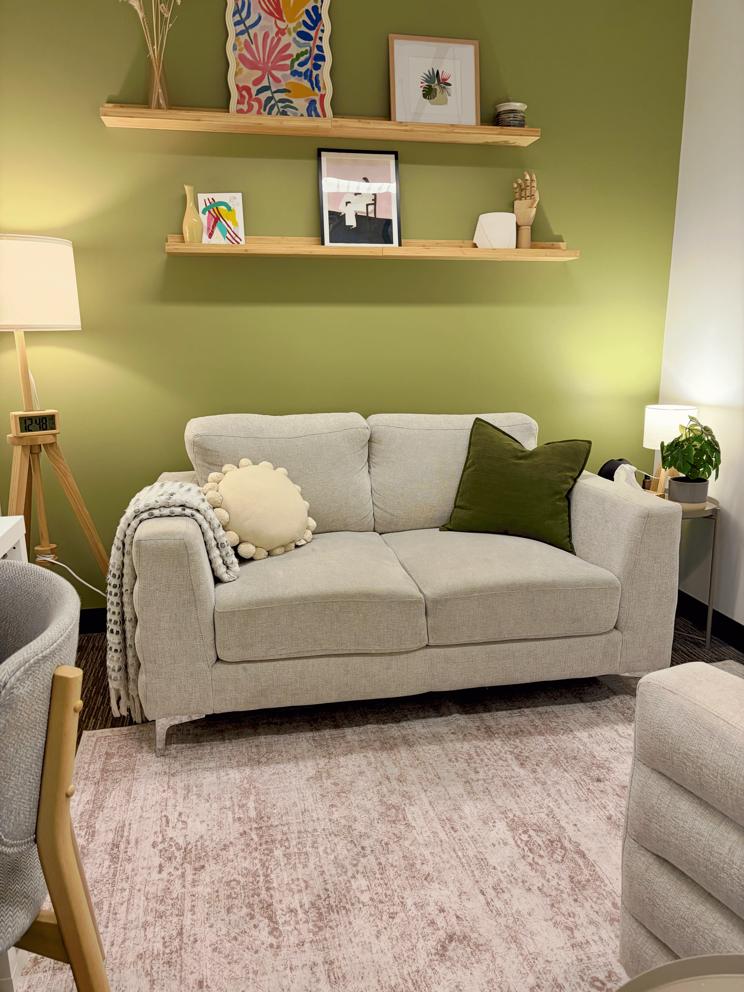 Living room with a beige sofa, green wall, and two wooden floating shelves with framed art and decorative items. A pink and beige patterned rug is on the floor. To the right, there's a bedside table with a lamp and a potted plant. To the left, a wooden floor lamp with a clock shows the time as 12:48.