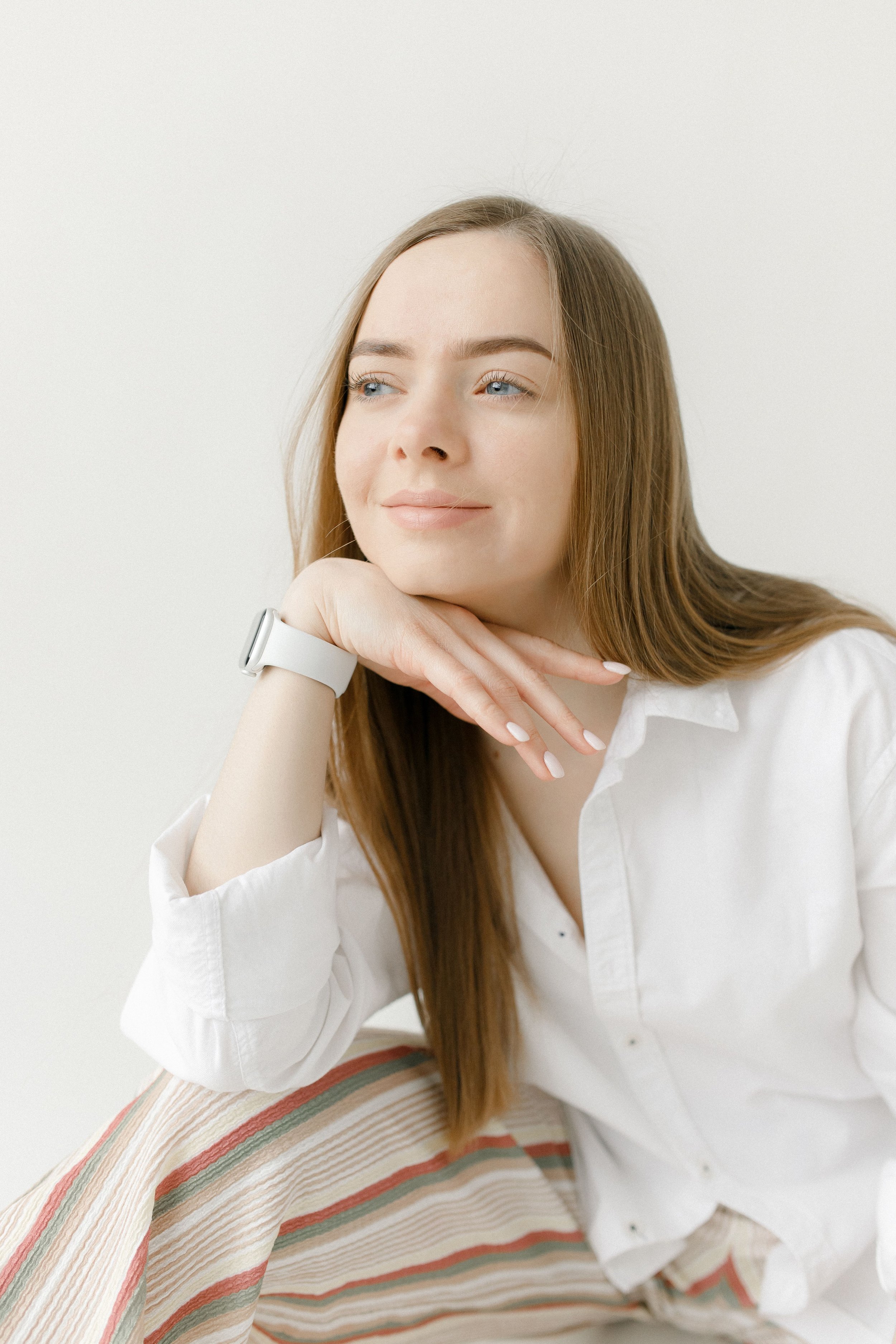 A young woman with long red hair and blue eyes, wearing a white shirt, rests her chin on her hand and looks slightly off to the side, sitting on a striped chair with a colorful pattern.