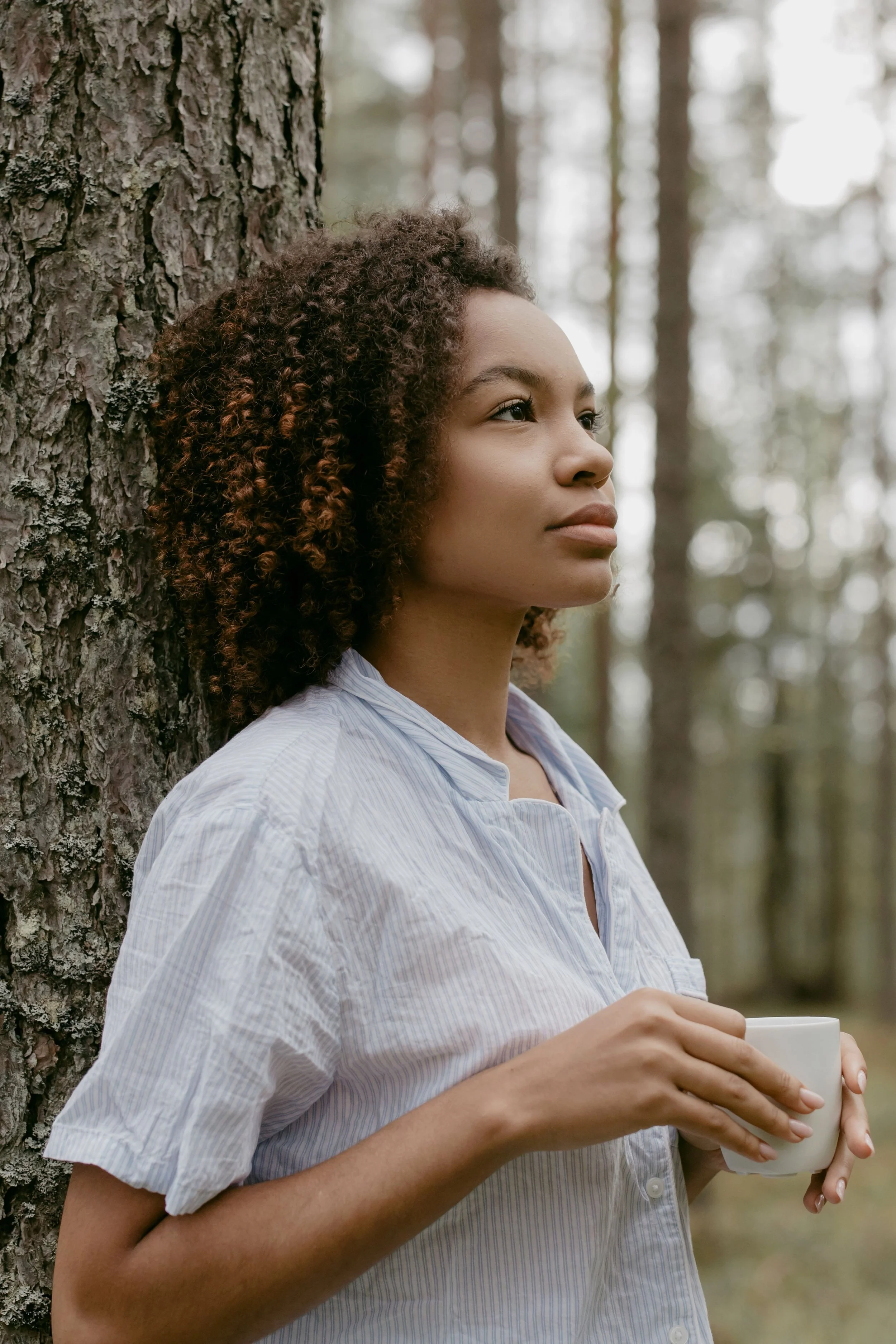 Young woman with curly hair holding a white mug leaning against a tree in a forest.