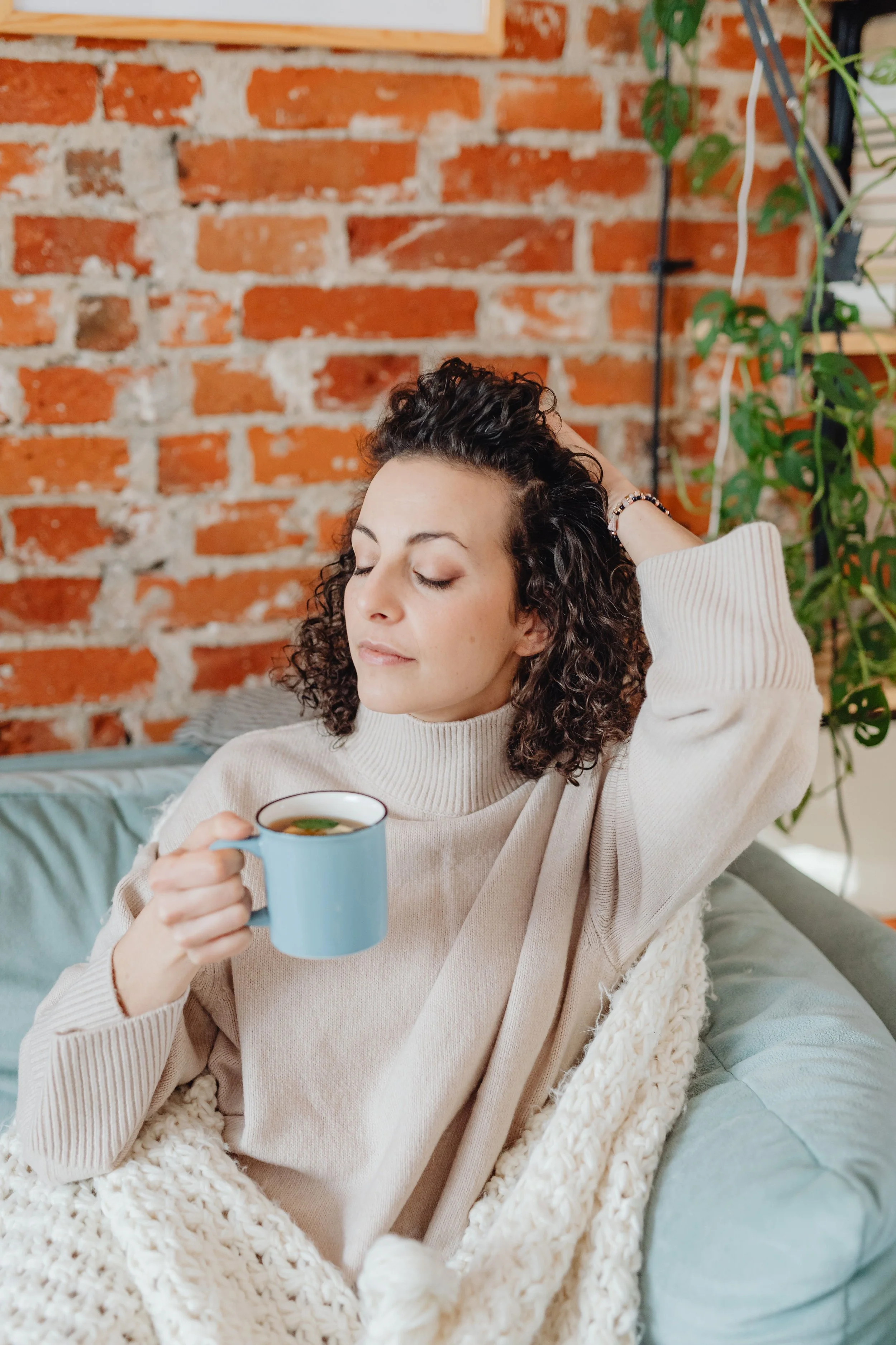 A woman with curly hair sitting on a couch, holding a blue mug, with a relaxed expression, wrapped in a chunky knit blanket, in a cozy room with a brick wall and green plants.