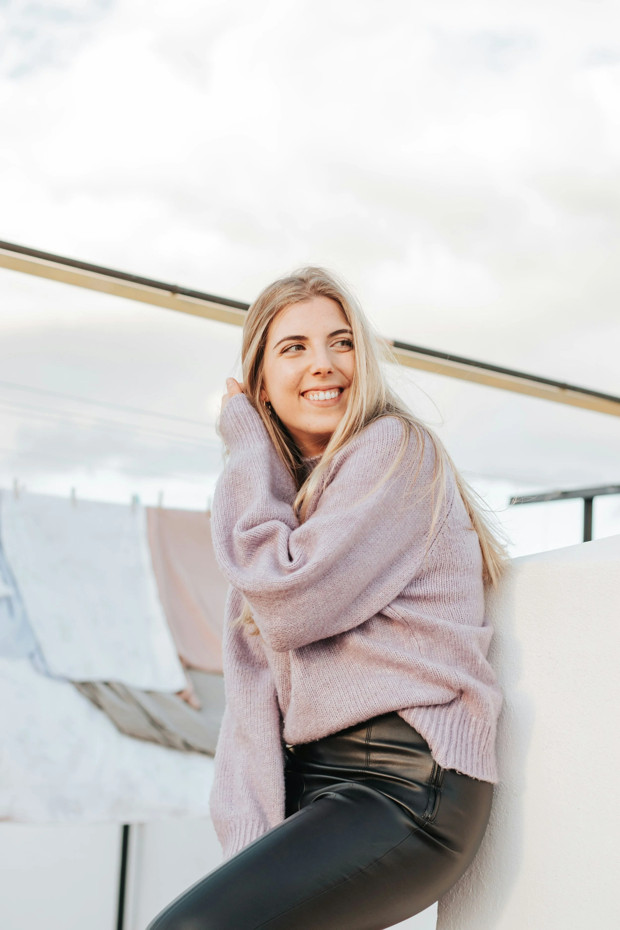 A young woman with long blonde hair smiling and resting her head on her hand, sitting outdoors with a gray sky in the background.