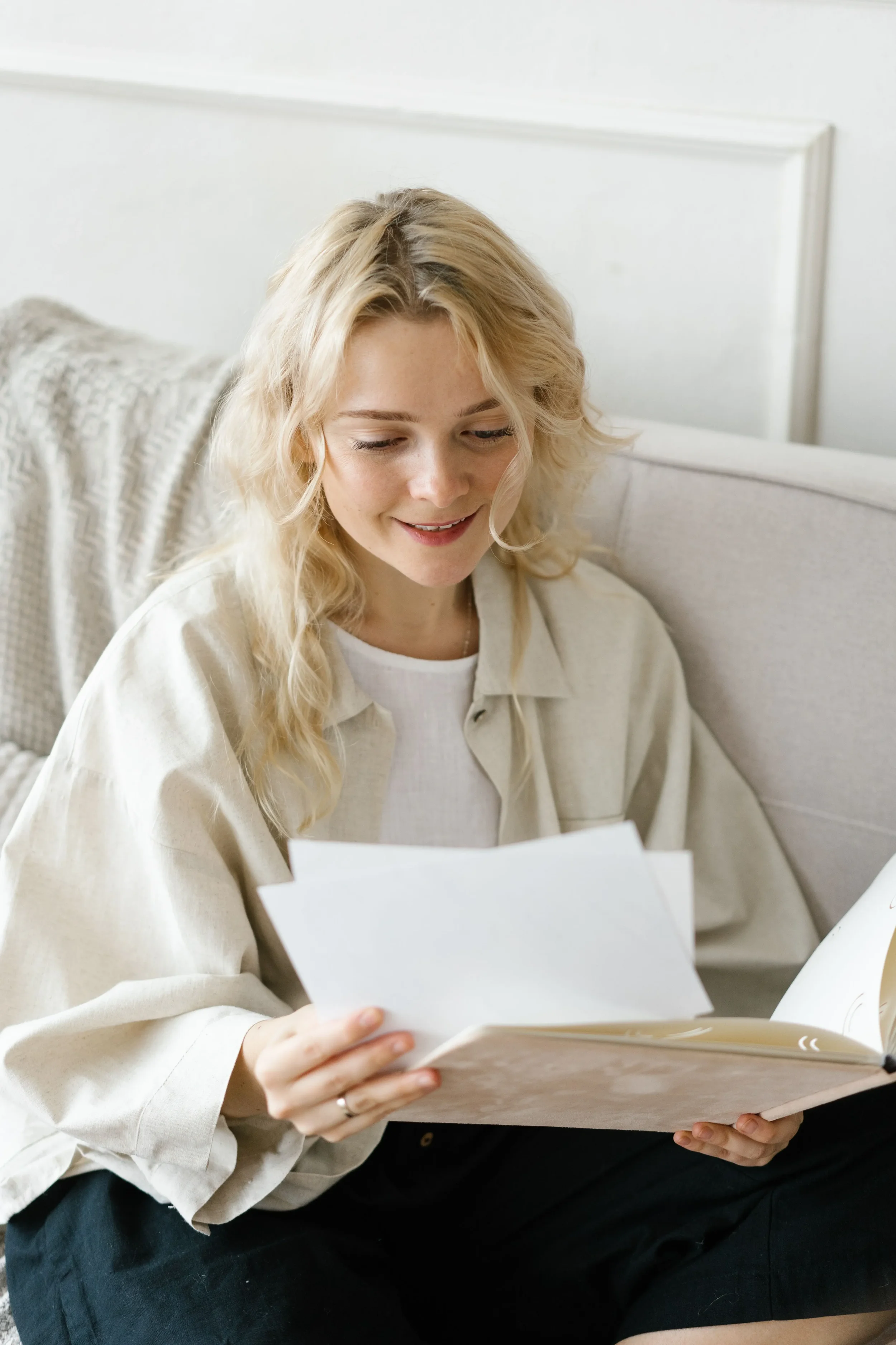 A young woman with blonde curly hair reading a book while sitting on a sofa, wearing a beige shirt and black shorts in a cozy, well-lit room.