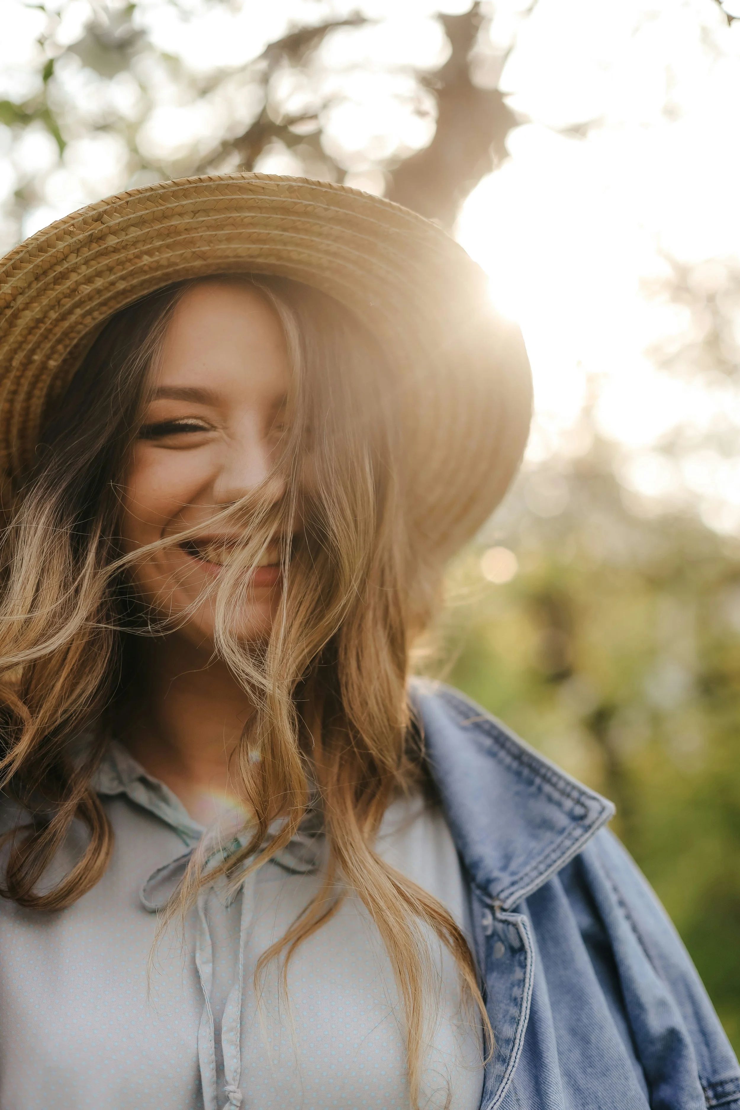 A young woman smiling outdoors wearing a wide-brimmed straw hat, denim jacket, and a light-colored blouse, with sunlight behind her.