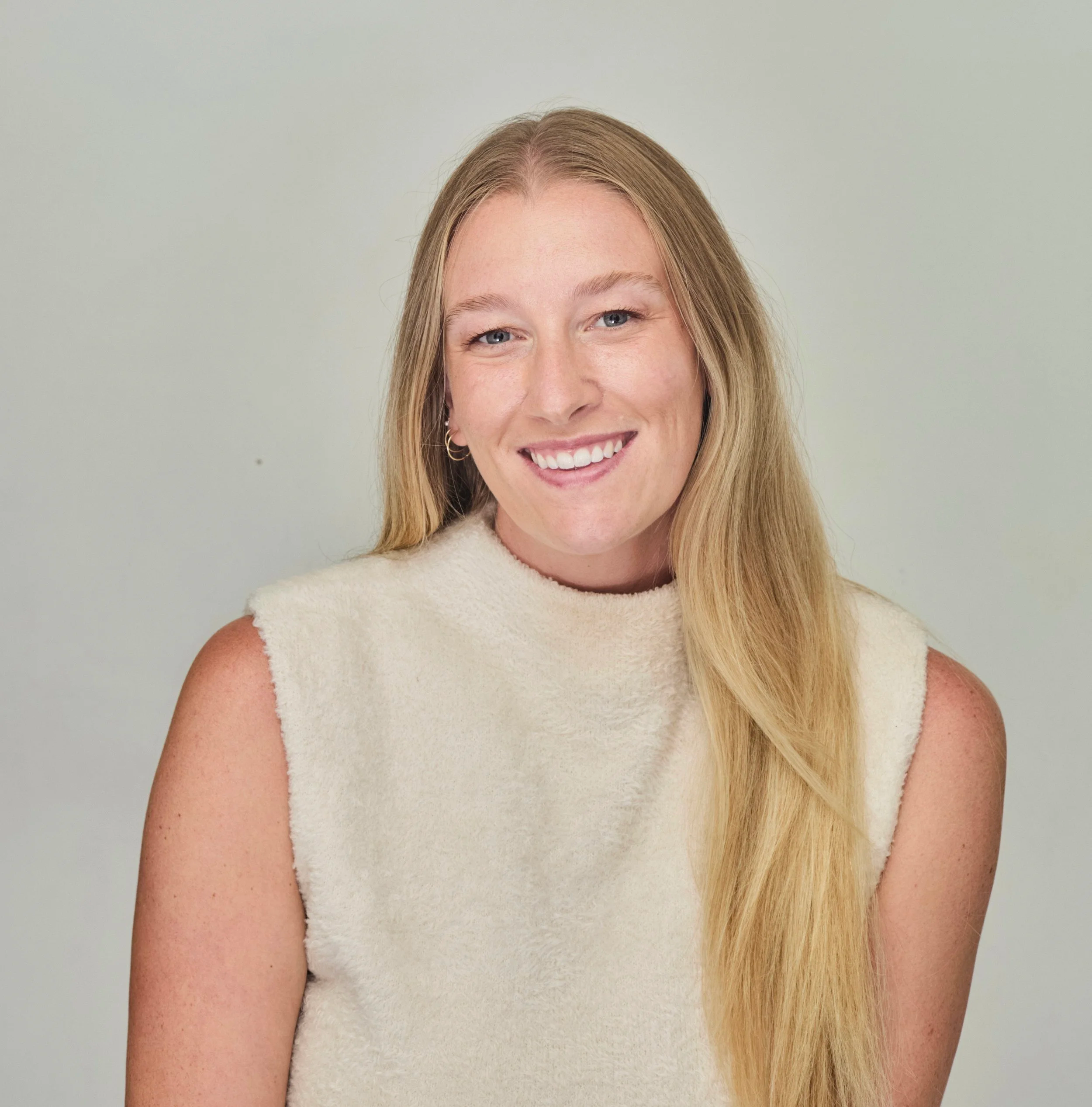 A young woman with long blonde hair smiling, wearing a sleeveless cream-colored top, standing against a plain light background.