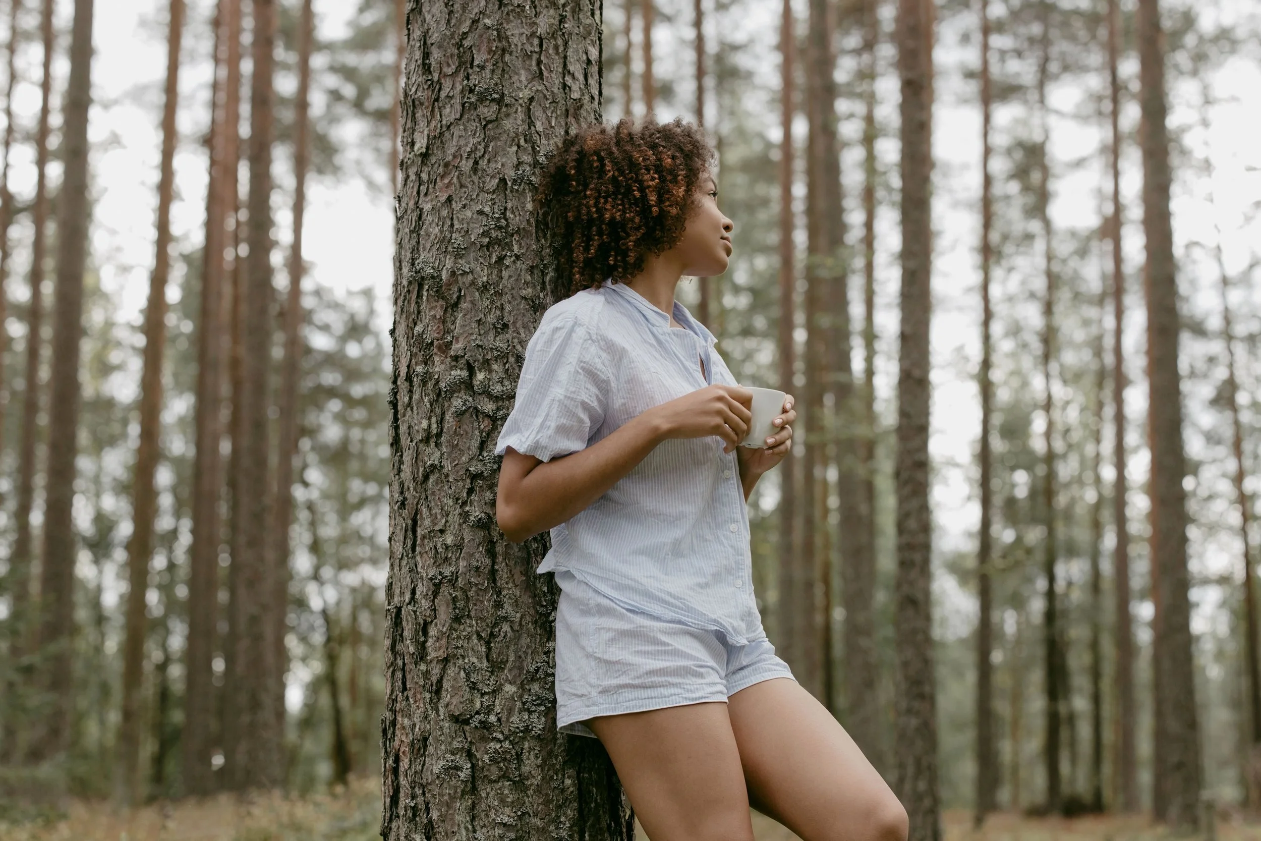 A woman with curly hair is leaning against a tree in a forest, holding a white mug, wearing a striped shirt and shorts, with a calm expression.