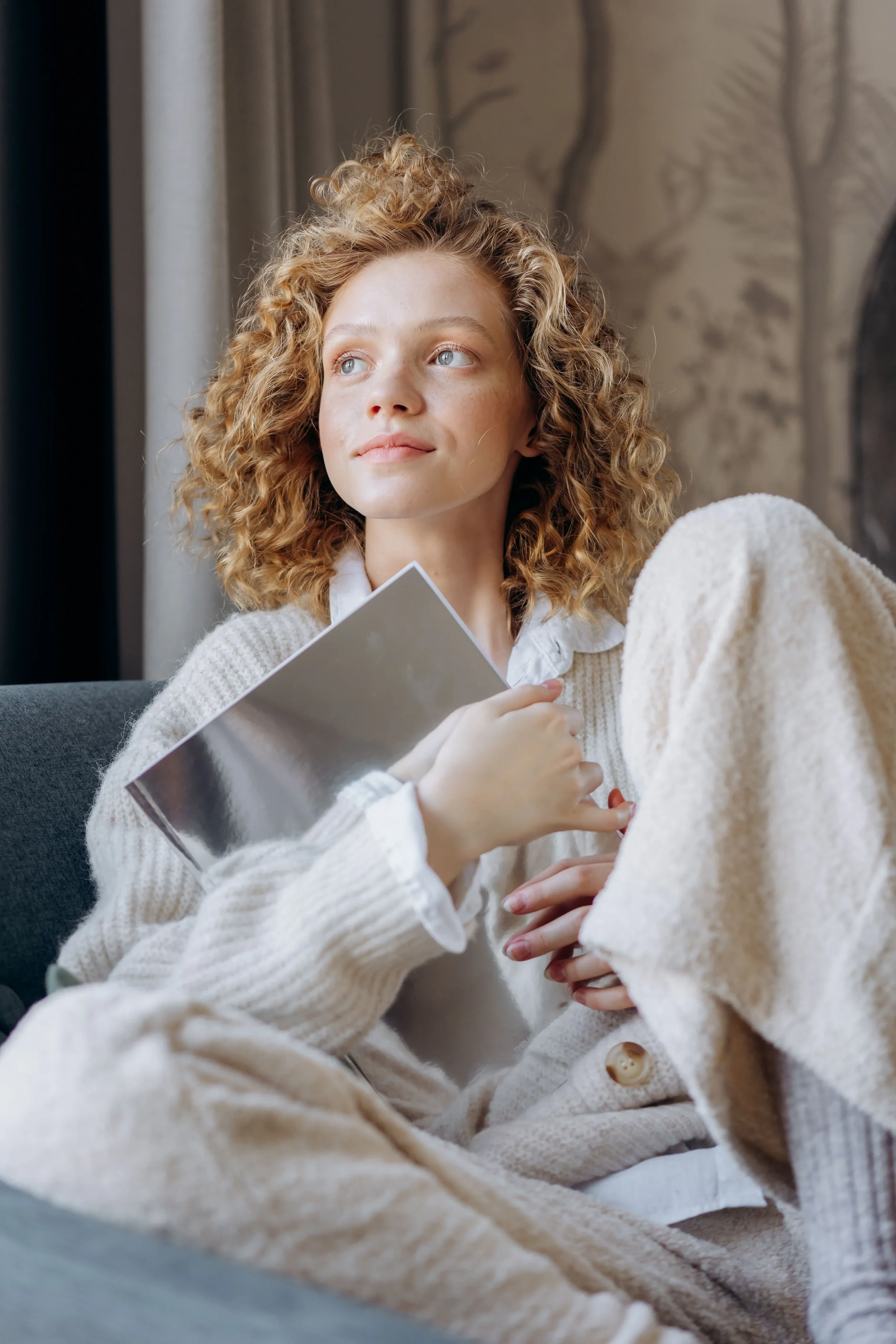 A woman with curly blonde hair and blue eyes sitting indoors near a window, holding a book and looking out thoughtfully.