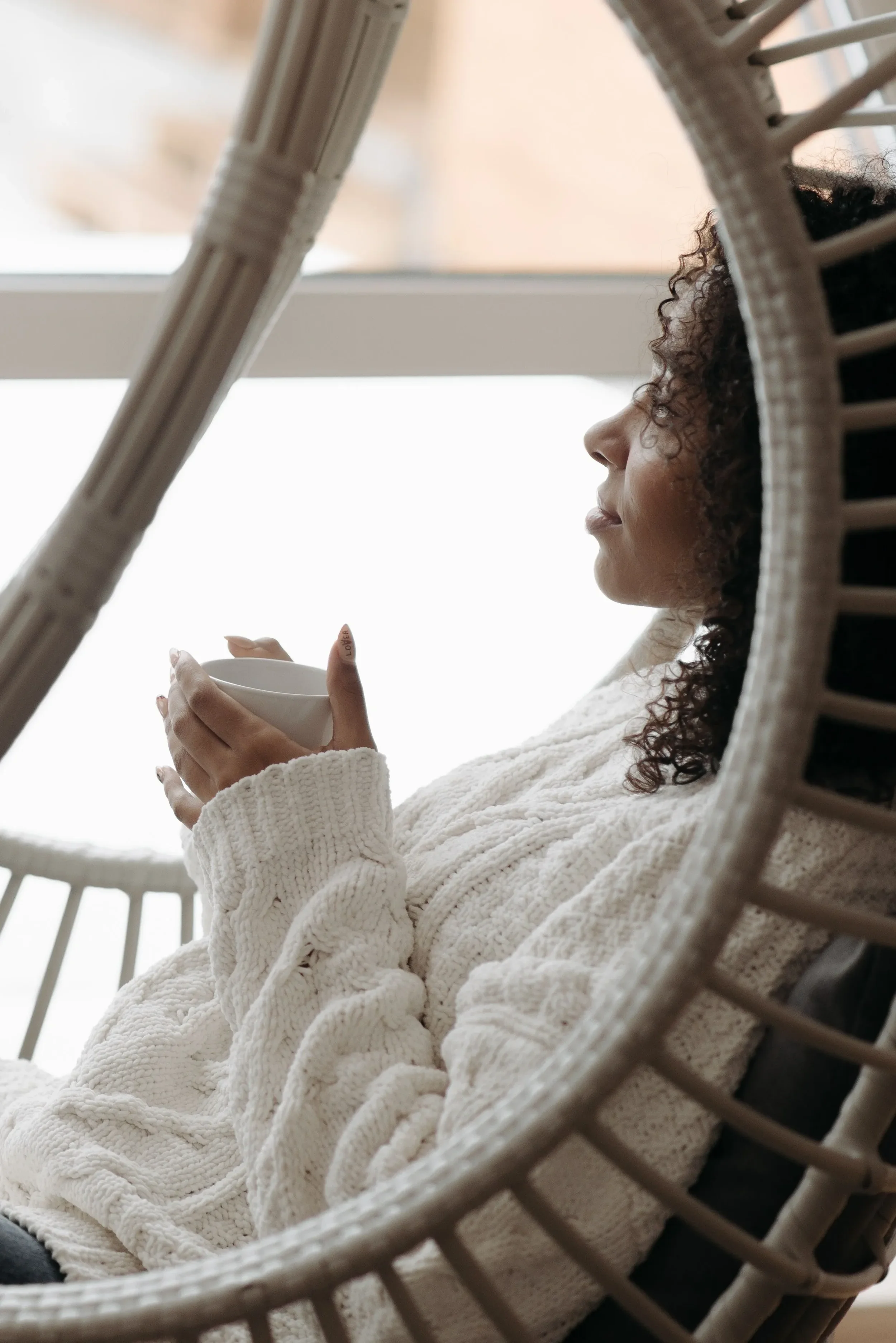 A woman with curly hair wearing a white chunky knit sweater, sitting in a circular wicker chair, holding a small white cup, looking out a window.
