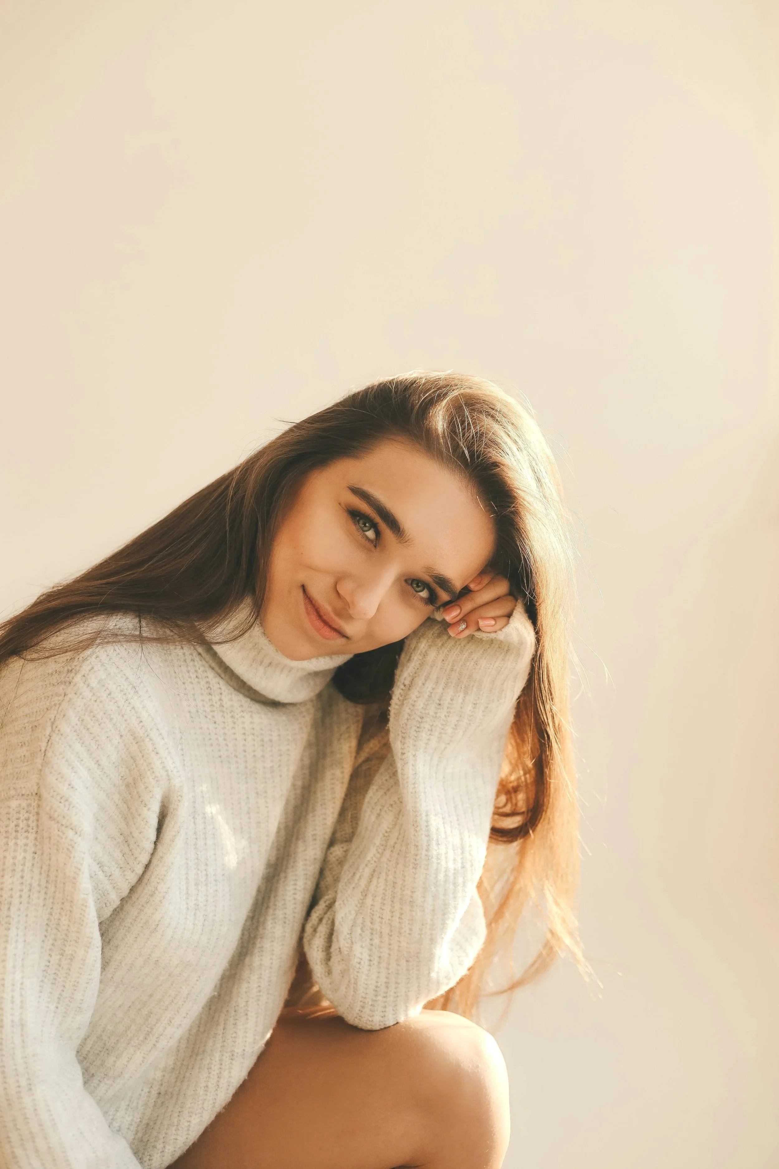 A young woman with long brown hair and green eyes, wearing a cream-colored sweater, rests her head on her hand and smiles at the camera, sitting against a plain light-colored background.