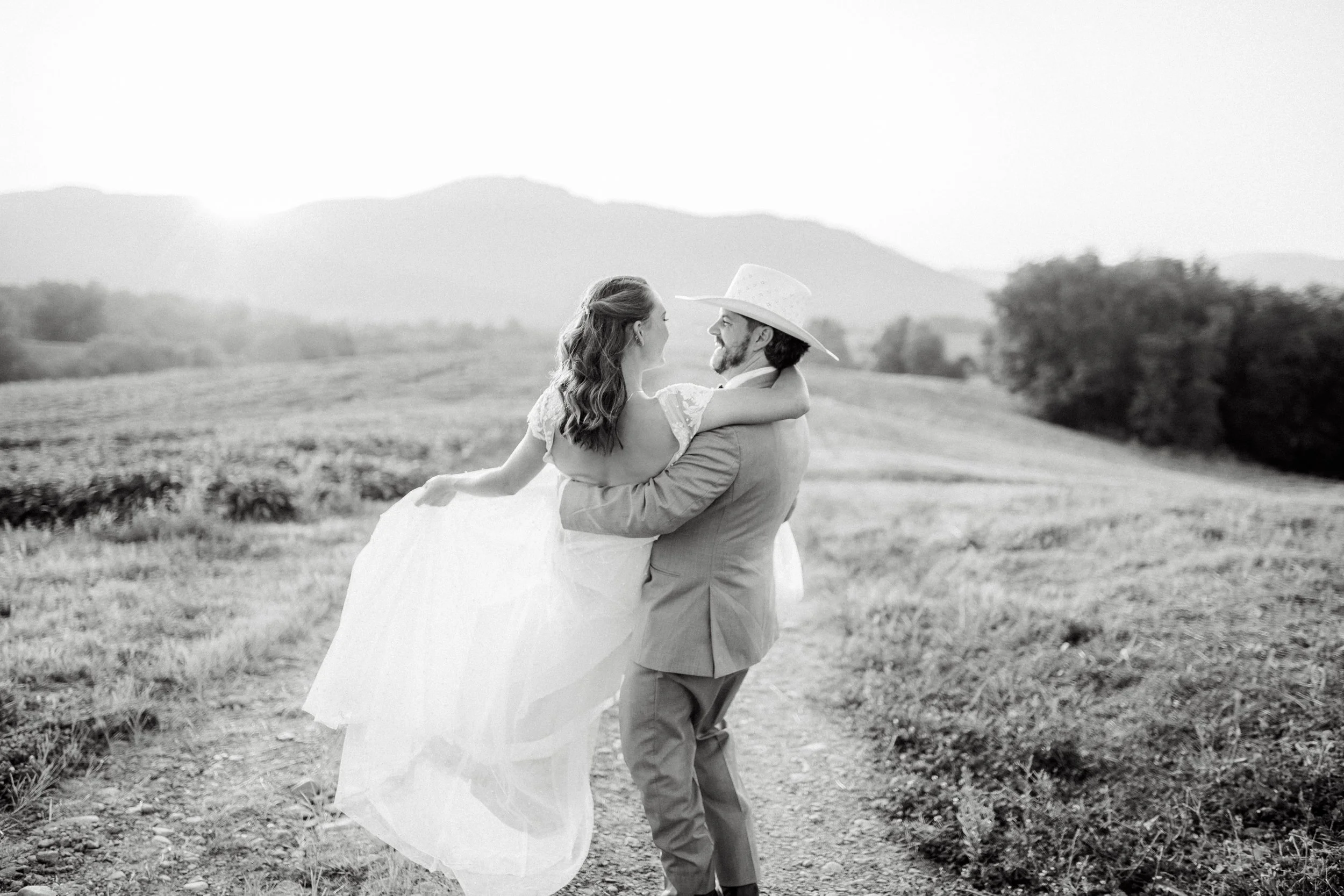 A couple in wedding attire, with the woman in a white wedding dress and the man in a suit with a wide-brimmed hat, embracing on a rural path with mountains in the background.