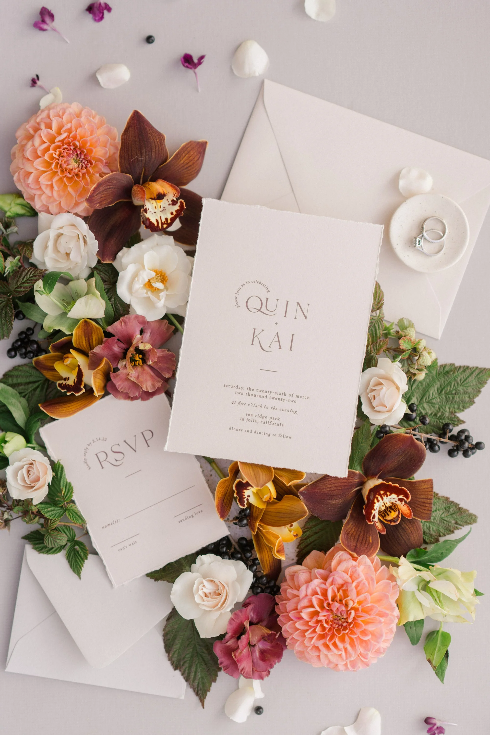 Wedding invitation surrounded by peach, white, and purple flowers, green leaves, and scattered petals, with white envelopes and a ring on a small white dish.