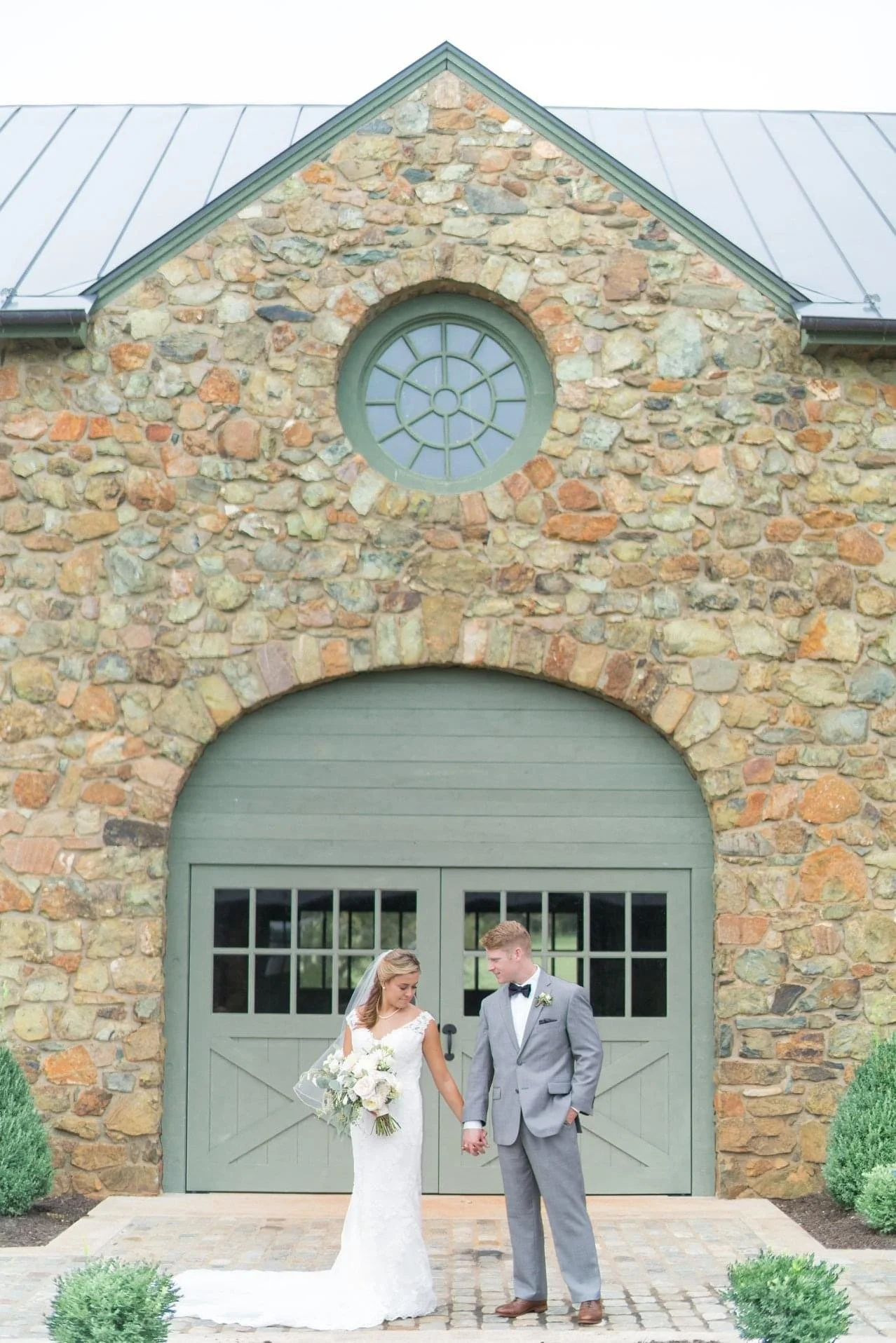 A bride and groom stand holding hands in front of a stone barn with green doors and a round window above, surrounded by small green bushes.