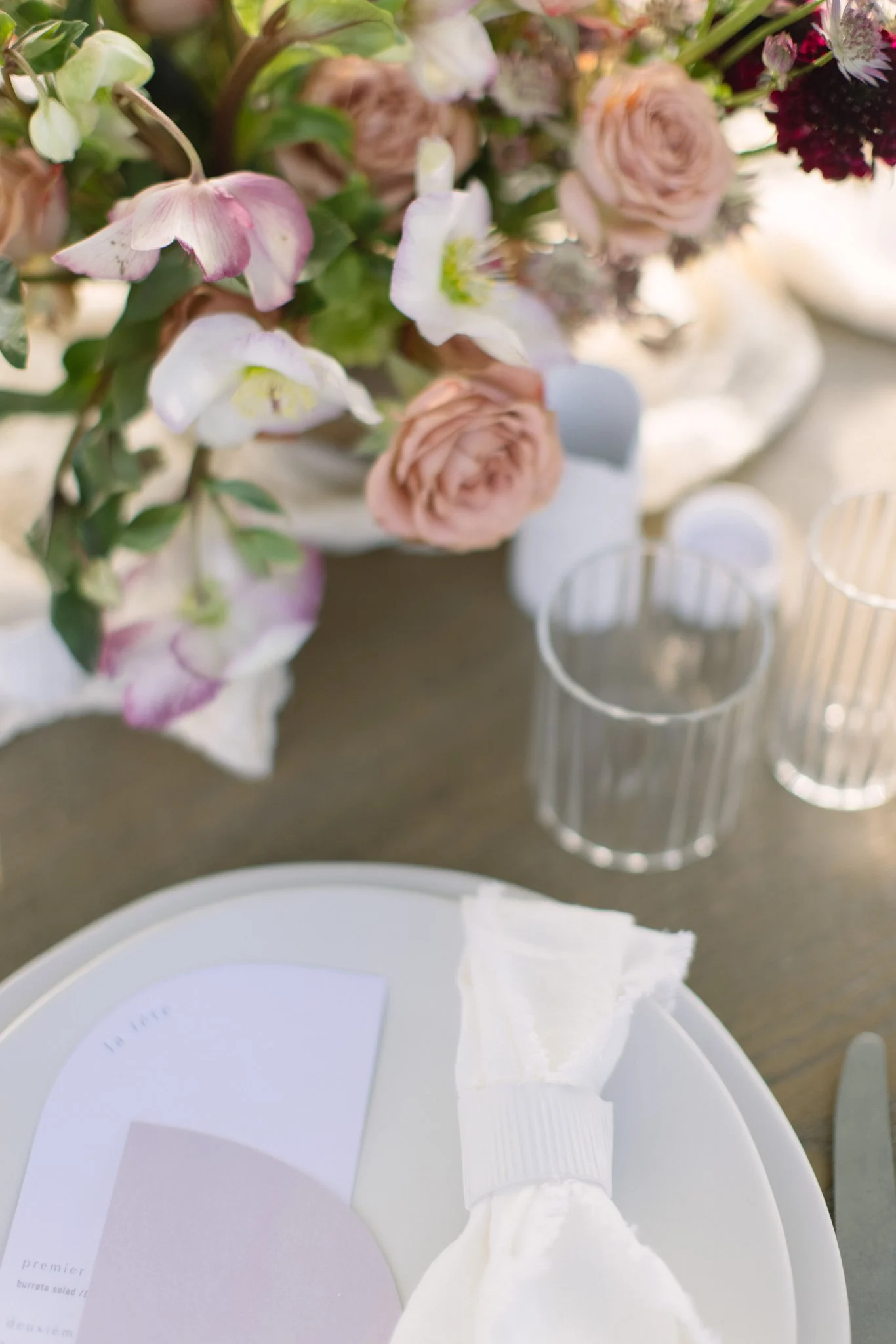 Close-up of a floral table centerpiece with pink, white, and purple flowers, glassware, and a place setting with a color palette card and napkin.