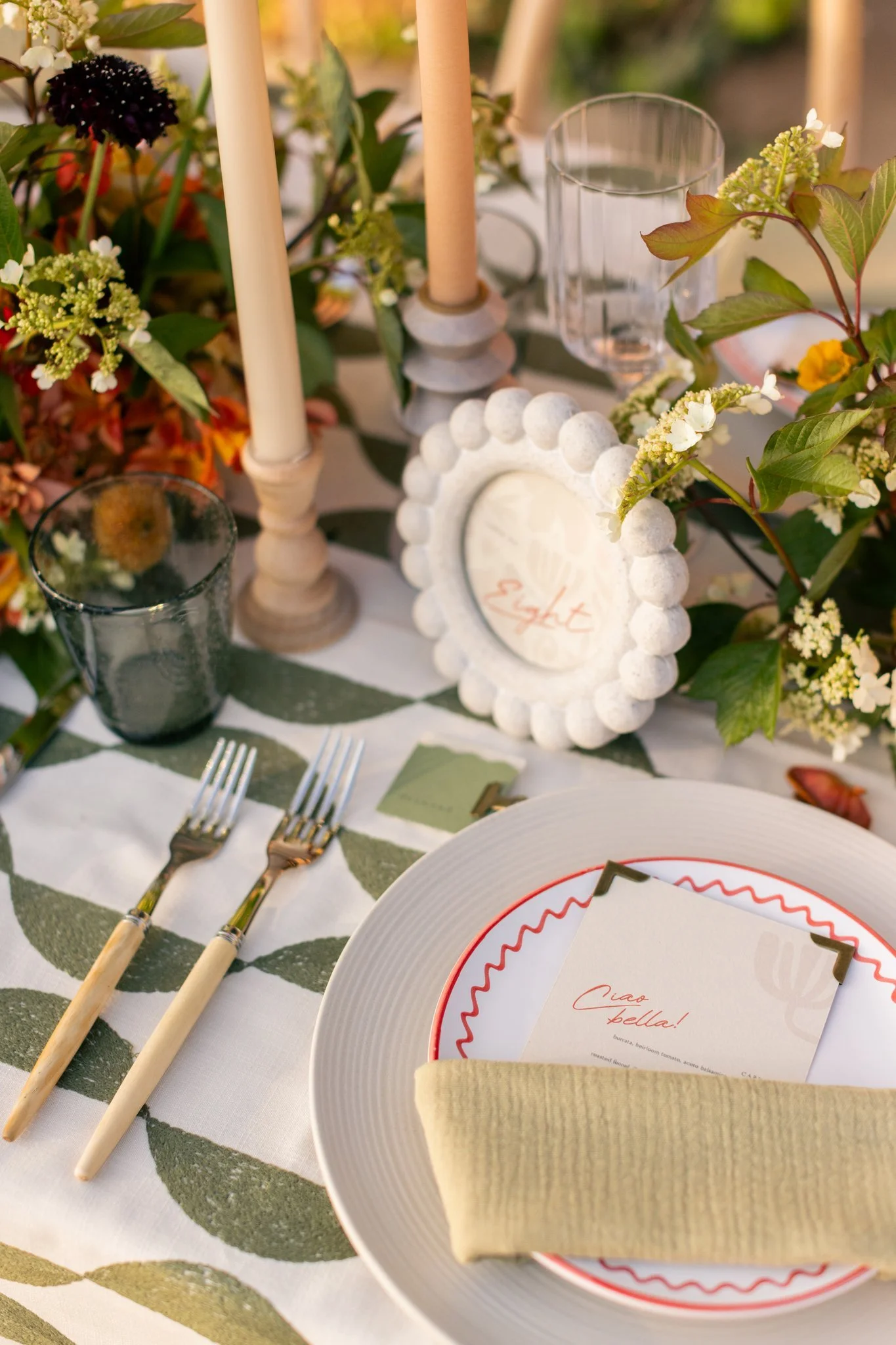 A beautifully set outdoor dining table with floral arrangements, candles, and a place setting with a napkin, plate, and menu card, decorated in a summery, elegant style.