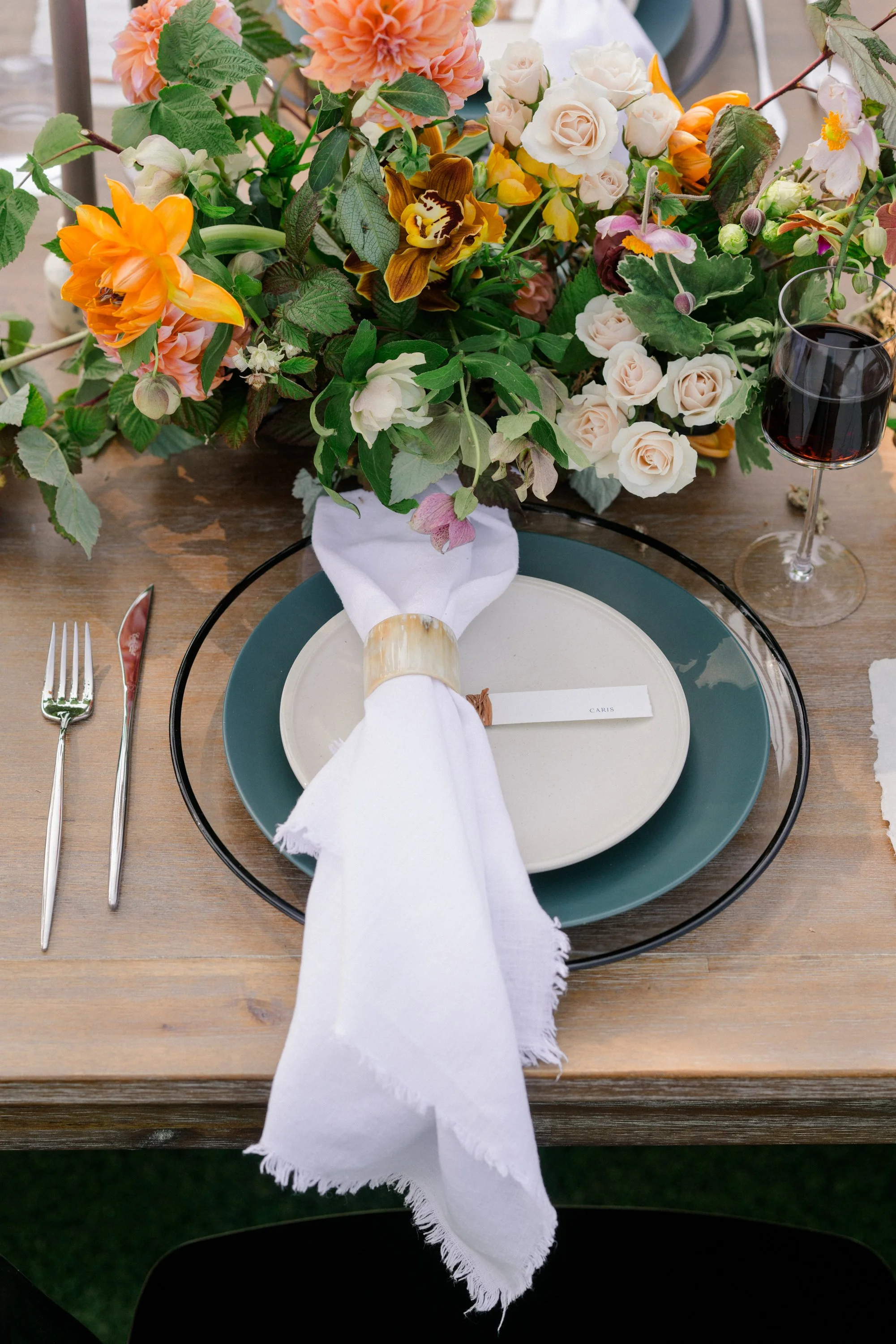 A table setting with a large floral centerpiece, place setting with a white napkin tied with a beige band, and a glass of red wine on a wooden table.