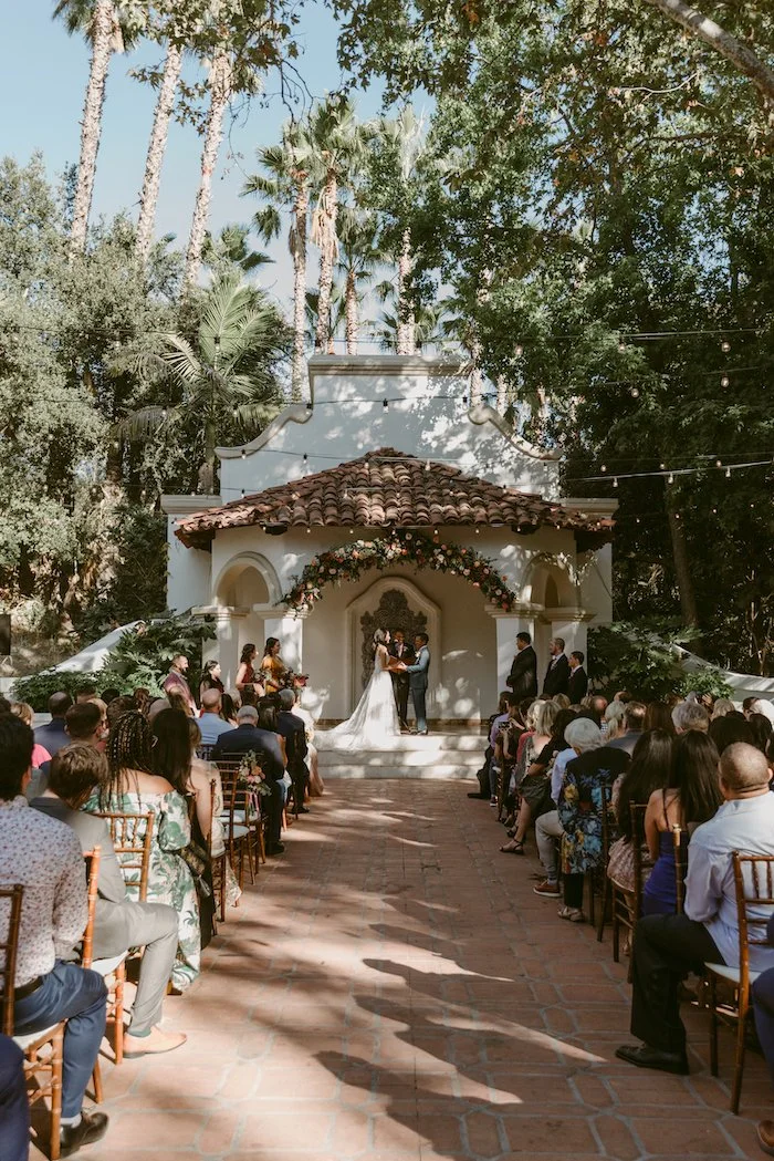 A wedding ceremony taking place outdoors in front of a small, white chapel with a tiled roof. The couple stands in front of an altar decorated with flowers, exchanging vows. Guests sit on wooden chairs, watching the ceremony surrounded by tall palm t