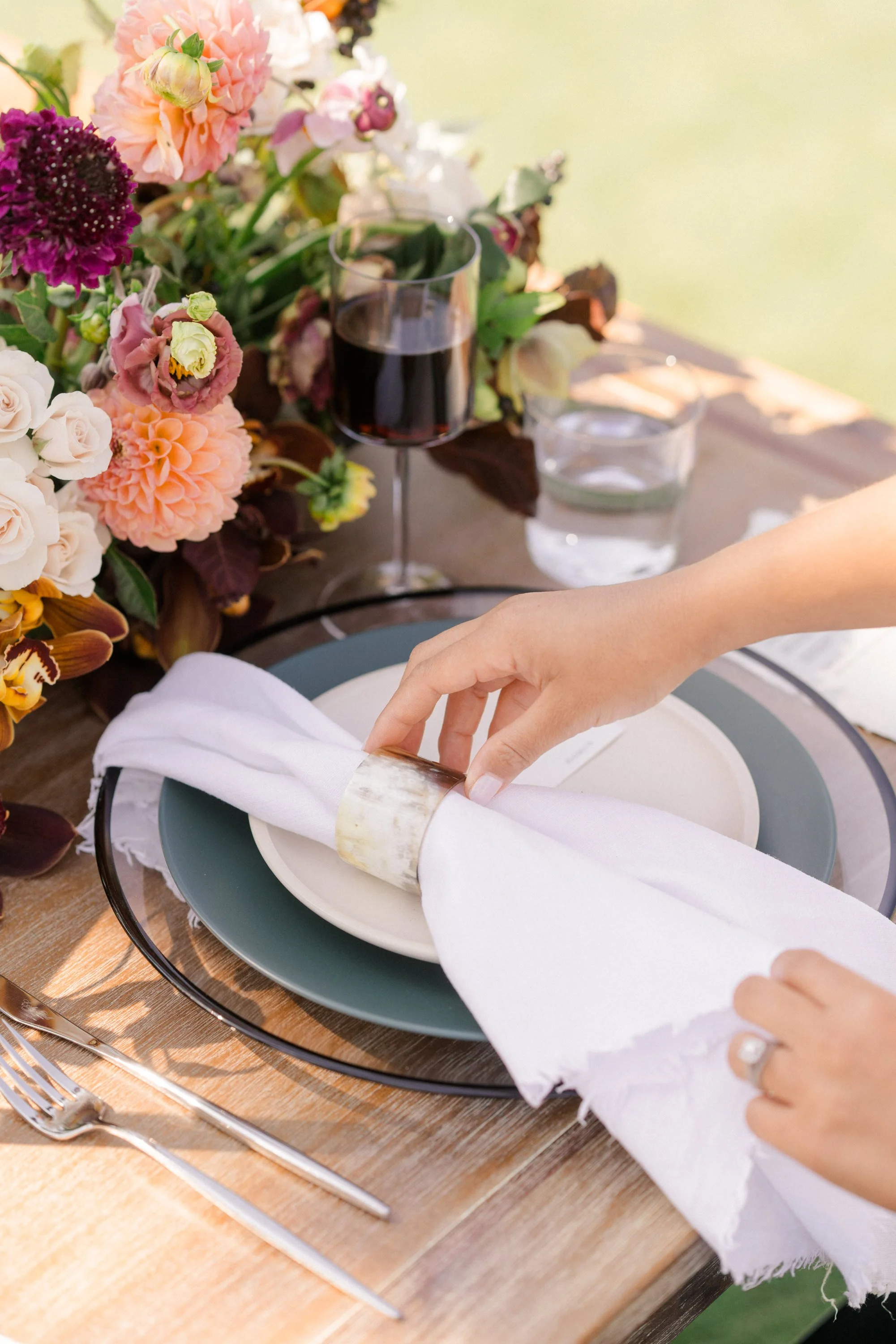 A person's hand adjusting a white napkin on a dinner plate, with a floral centerpiece, a glass of red wine, and a glass of water on a wooden table.