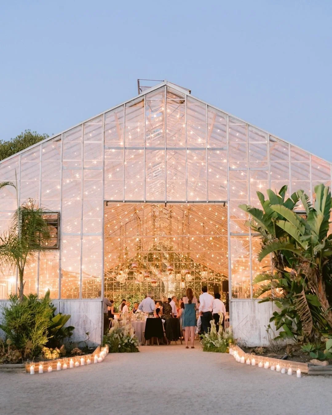 A glass greenhouse decorated with string lights, with people gathered inside for a celebration or event, surrounded by greenery and candles outside.