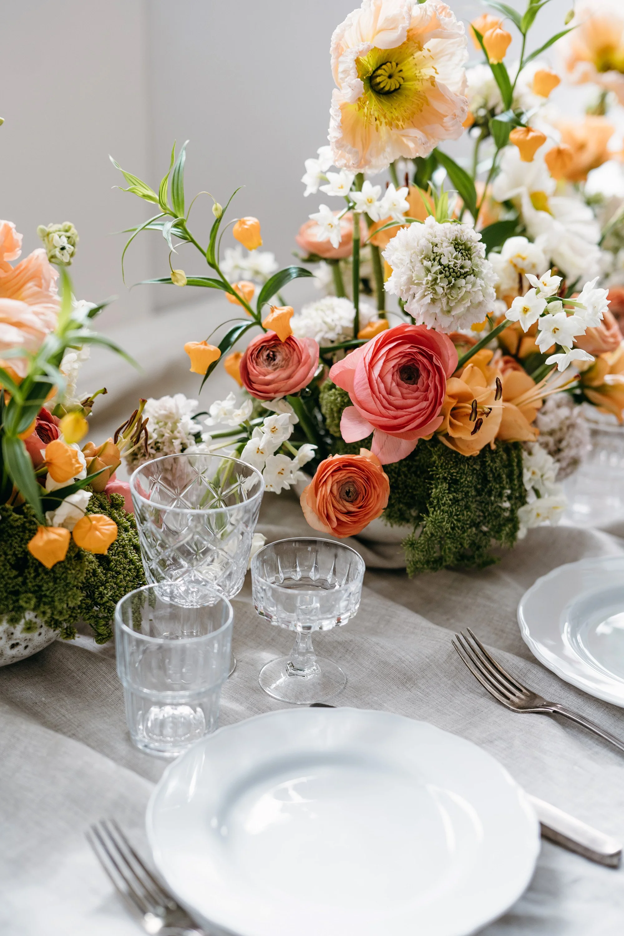 Elegant table setting with white plates, crystal glasses, and a large floral centerpiece of pink, white, and orange flowers.