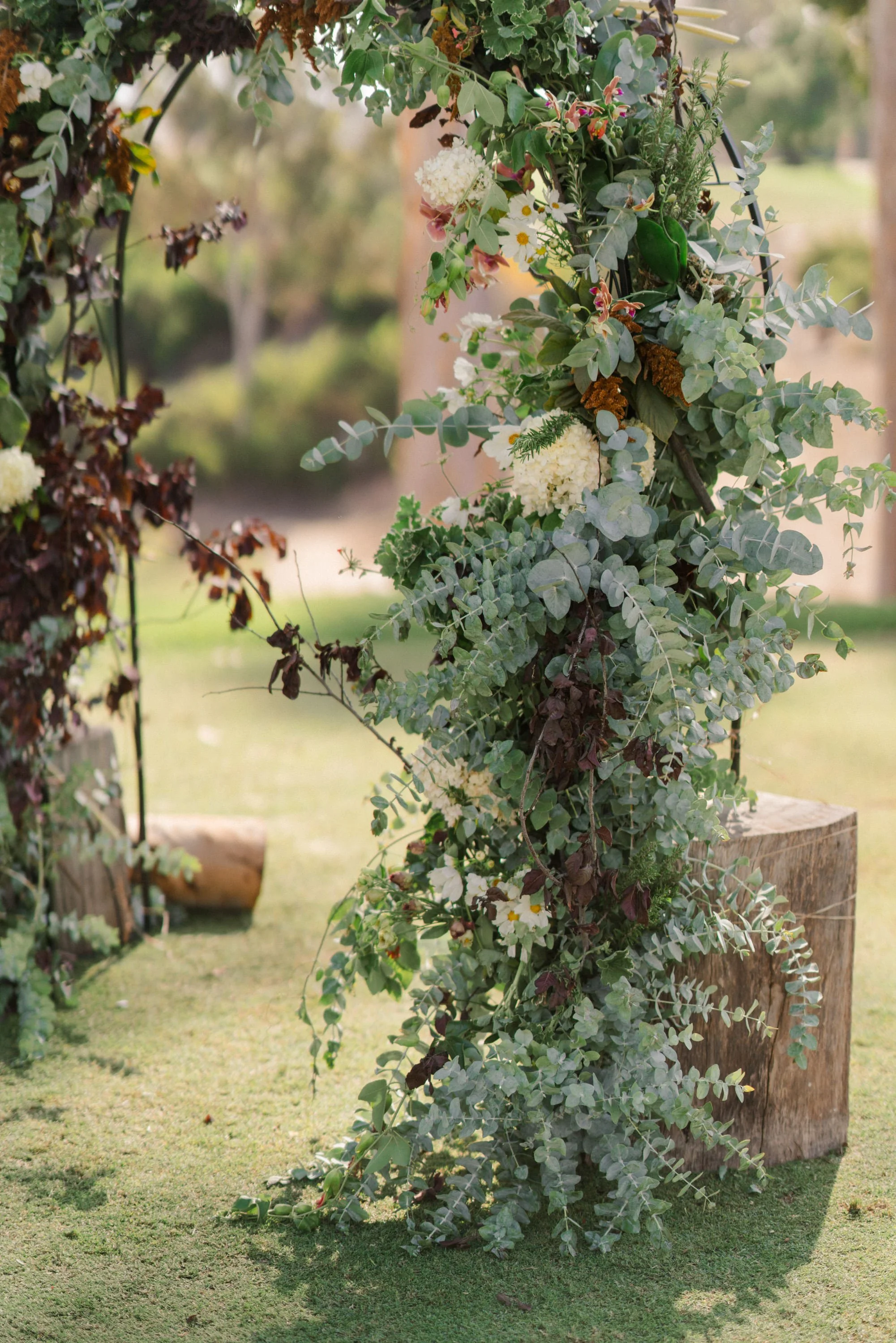 A floral arch decorated with various green foliage and white flowers, positioned on a grassy outdoor area.
