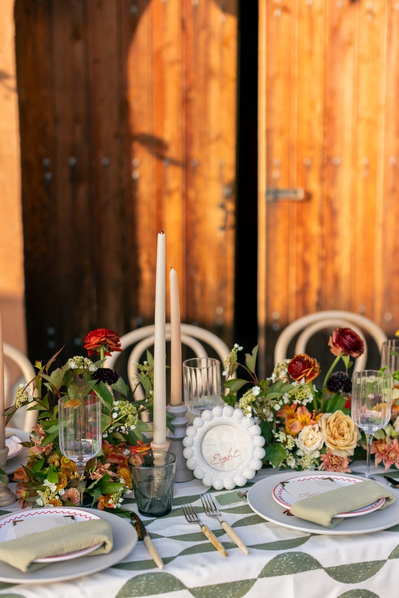 A decorated wedding or event table with floral arrangements, candles, glassware, plates, and cutlery, set outdoors against a wooden fence.