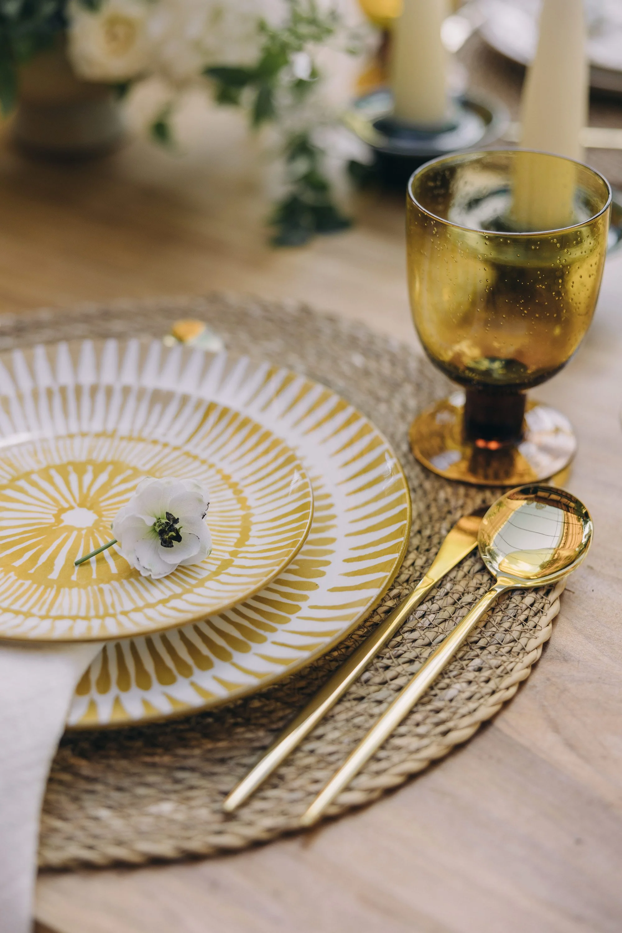 Elegant table setting with gold-rimmed yellow and white patterned plates, a white flower, gold utensils, a woven placemat, and a gold glass with a drink, on a wooden table.