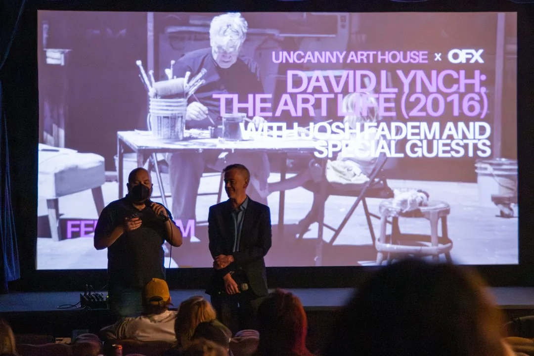 Two men standing in front of a movie screen that shows a still from David Lynch's The Art Life documentary