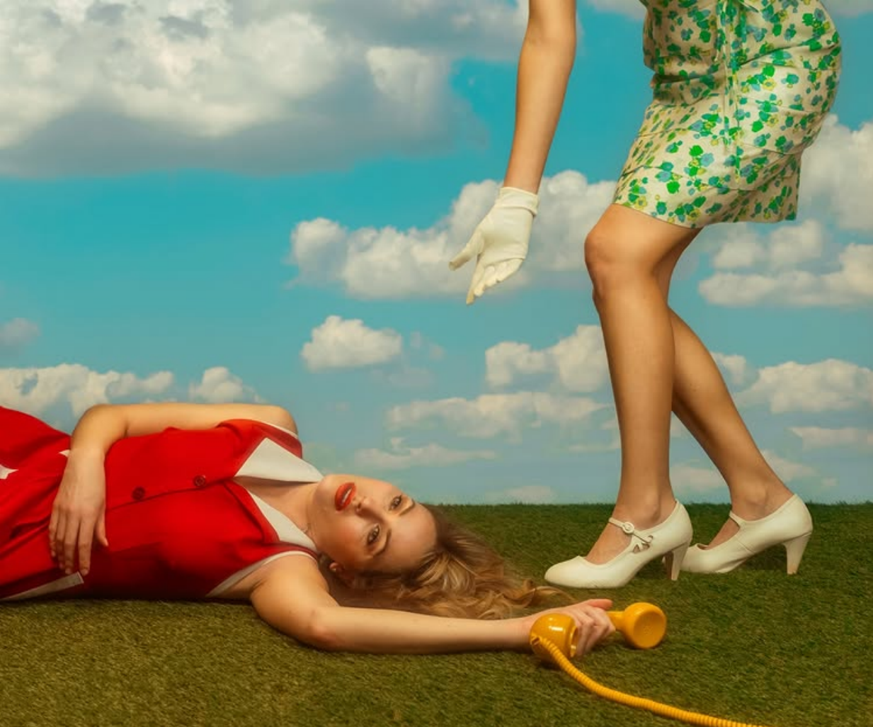 A woman lies on the grass in a red dress, holding a yellow phone in her hand while the bottom half of a woman in a green floral dress, wearing white shoes and a white glove reaches down to her