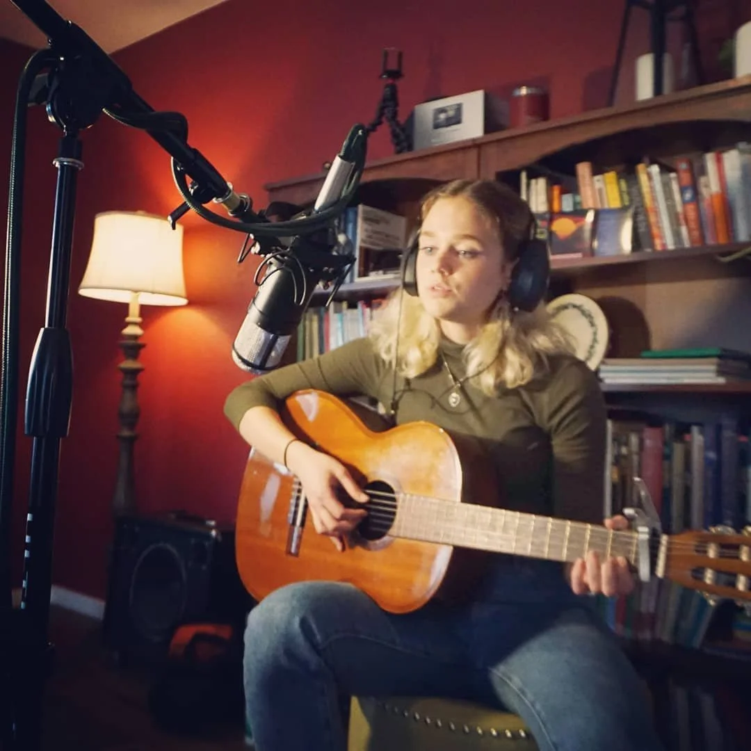 A girl sitting in front of a microphone with headphones on and holding a guitar inside of a bedroom with a book shelf behind her, red walls, and a lamp.